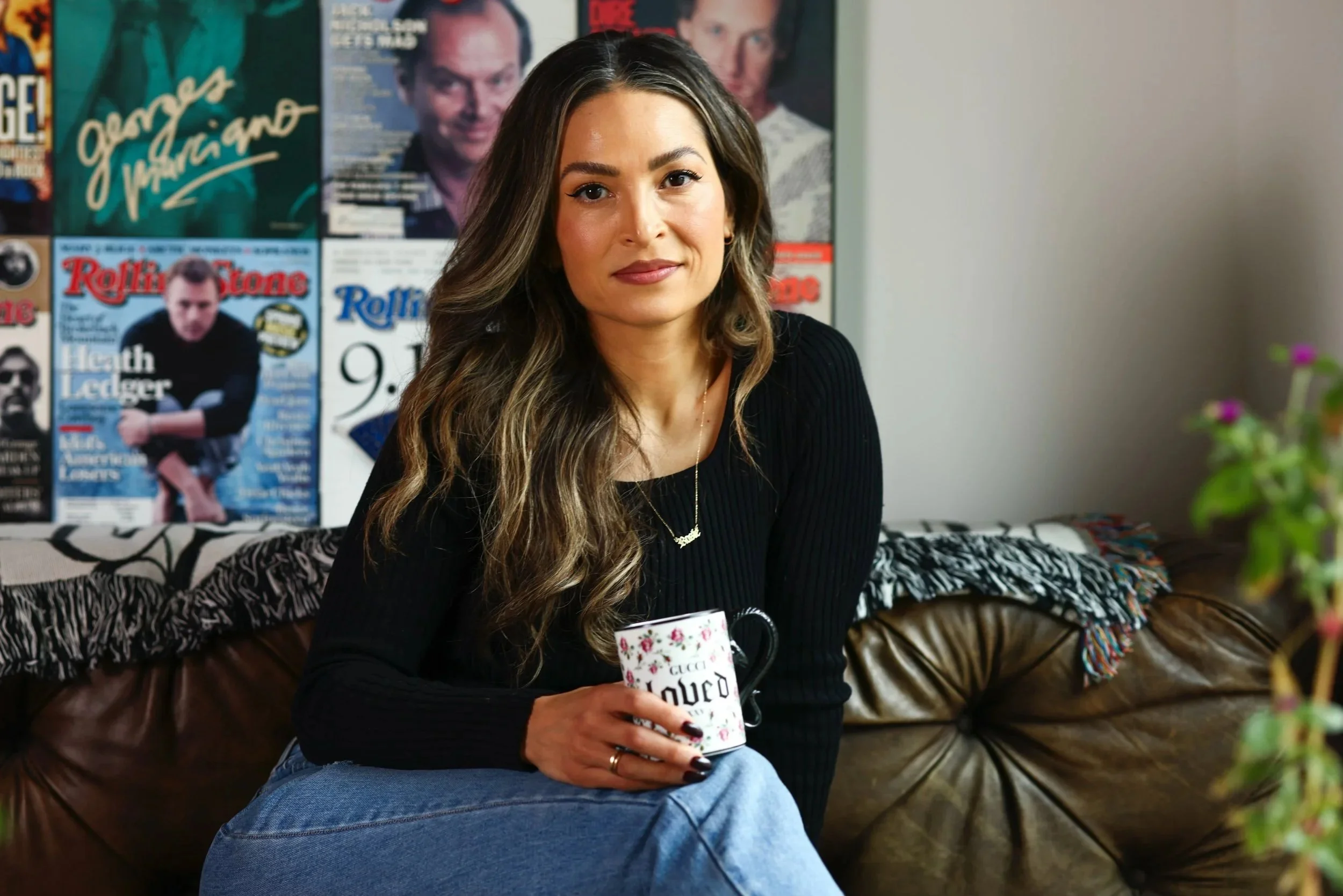 A woman with wavy brown hair and wearing a black top, holding a floral patterned mug, sitting on a leather couch in a room with a collage of magazine covers and posters on the wall behind her.