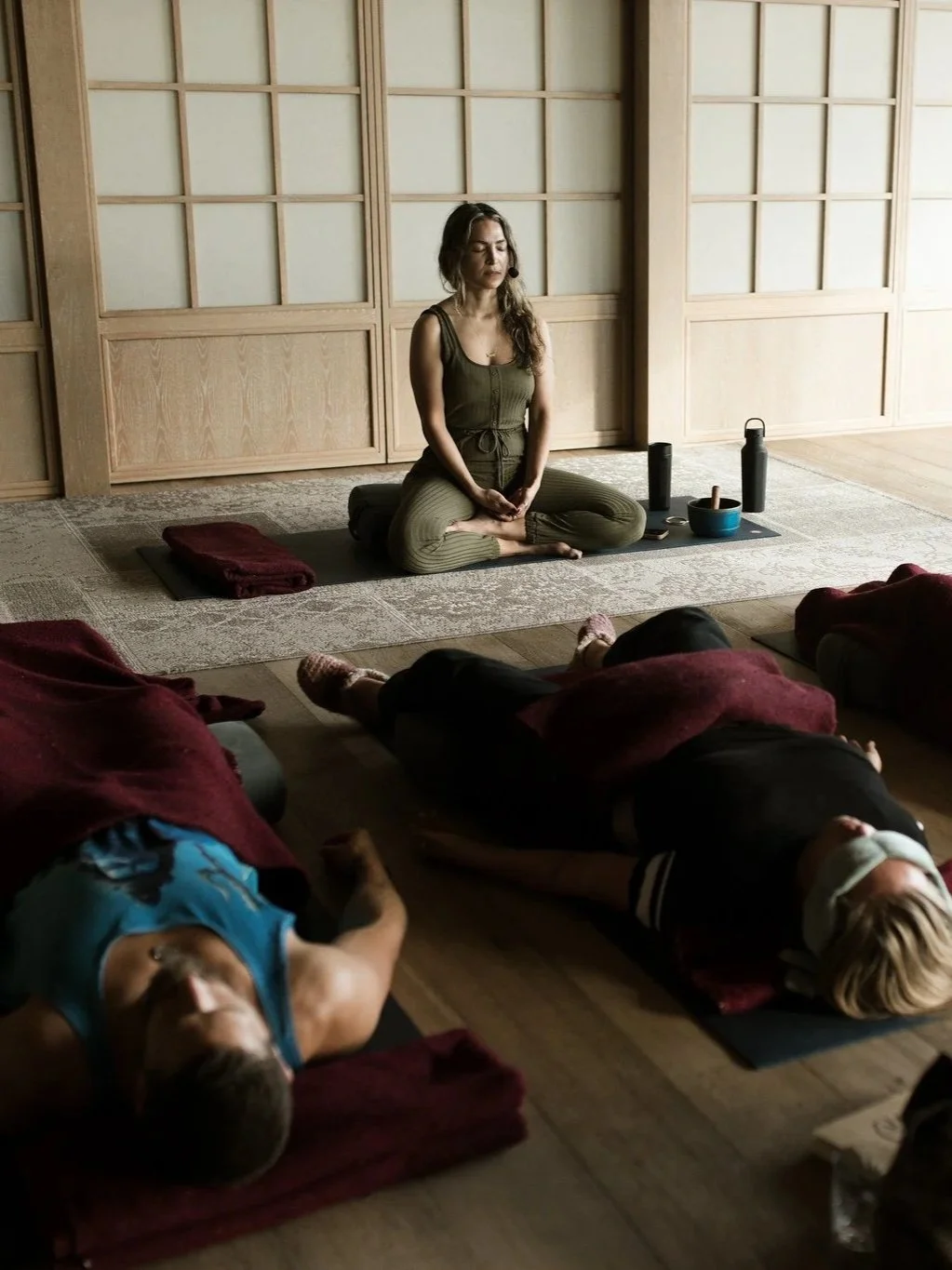Women practicing yoga in a peaceful studio with natural light, sitting in a cross-legged meditation pose at the front, with others lying down on mats.