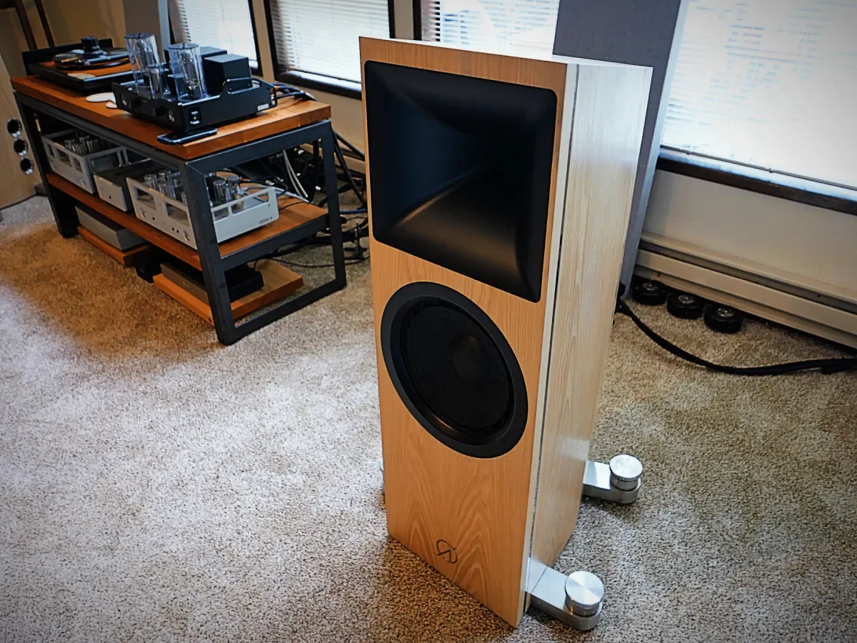 A large wooden floor-standing speaker with a black driver and horn, positioned in a room with beige carpet and a window. A metal equipment cart with audio and electronic gear is nearby.
