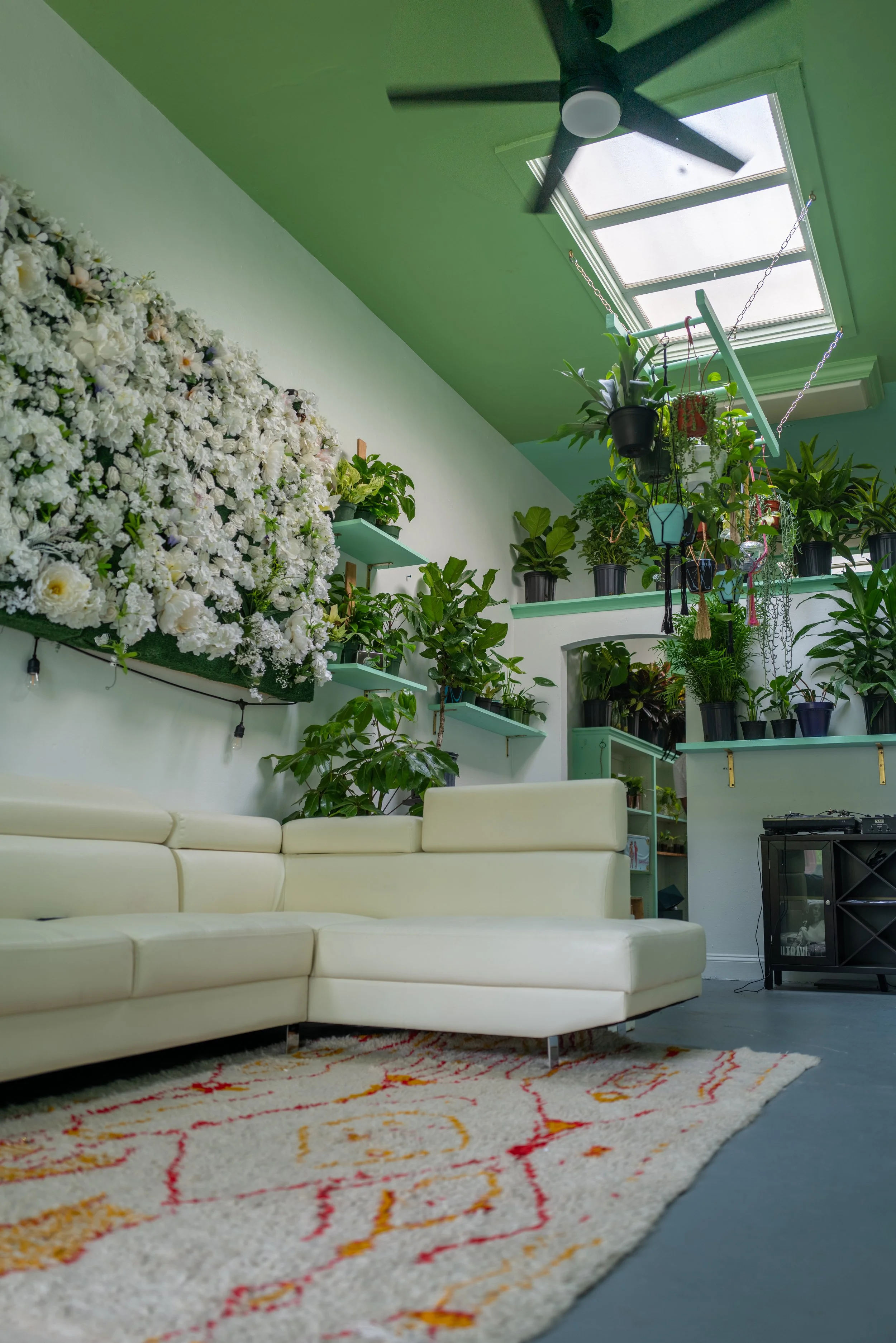 Living room with a white sectional sofa, a patterned rug, green walls, indoor plants, a ceiling fan, and a skylight.