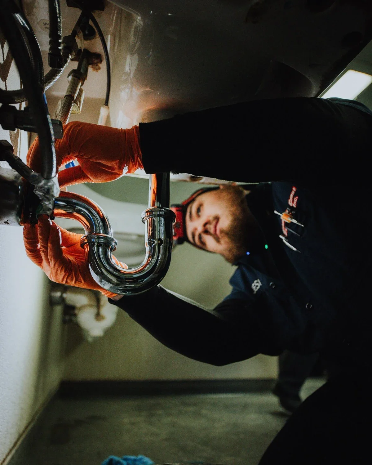 A technician wearing orange gloves working on plumbing pipes under a sink, smiling and looking at the pipes.