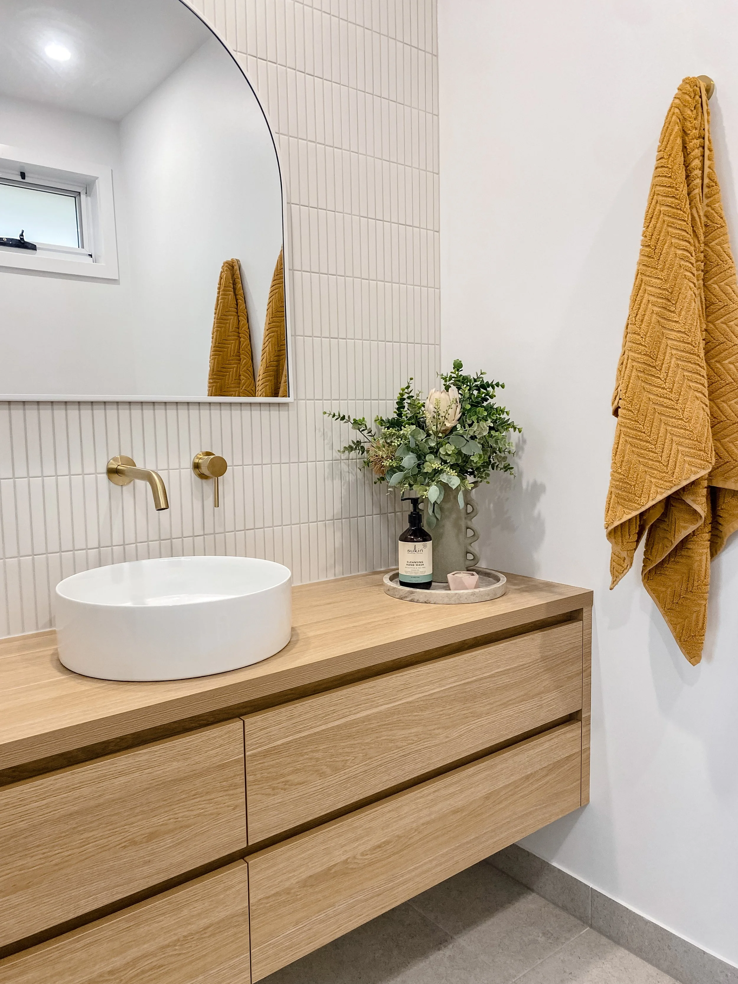 Modern bathroom vanity with a round white vessel sink, gold faucet, and a bouquet of green and white flowers in a black vase on a marble tray. There are two mustard yellow towels hanging on hooks and a small window in the background.