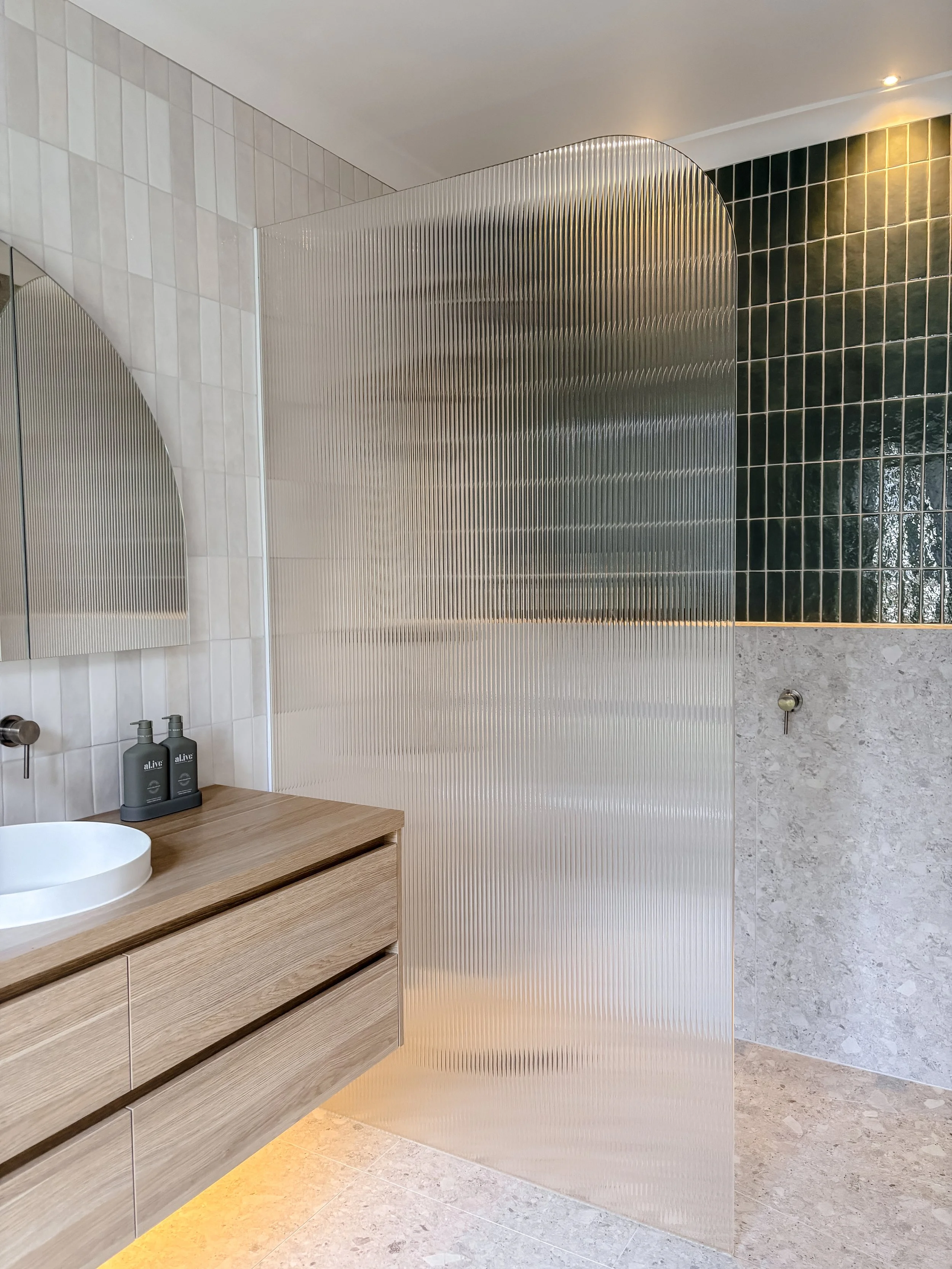 A modern bathroom with a wooden vanity, a white vessel sink, and two gray soap dispensers. There is a textured frosted glass partition and contrasting black and beige tiled walls.