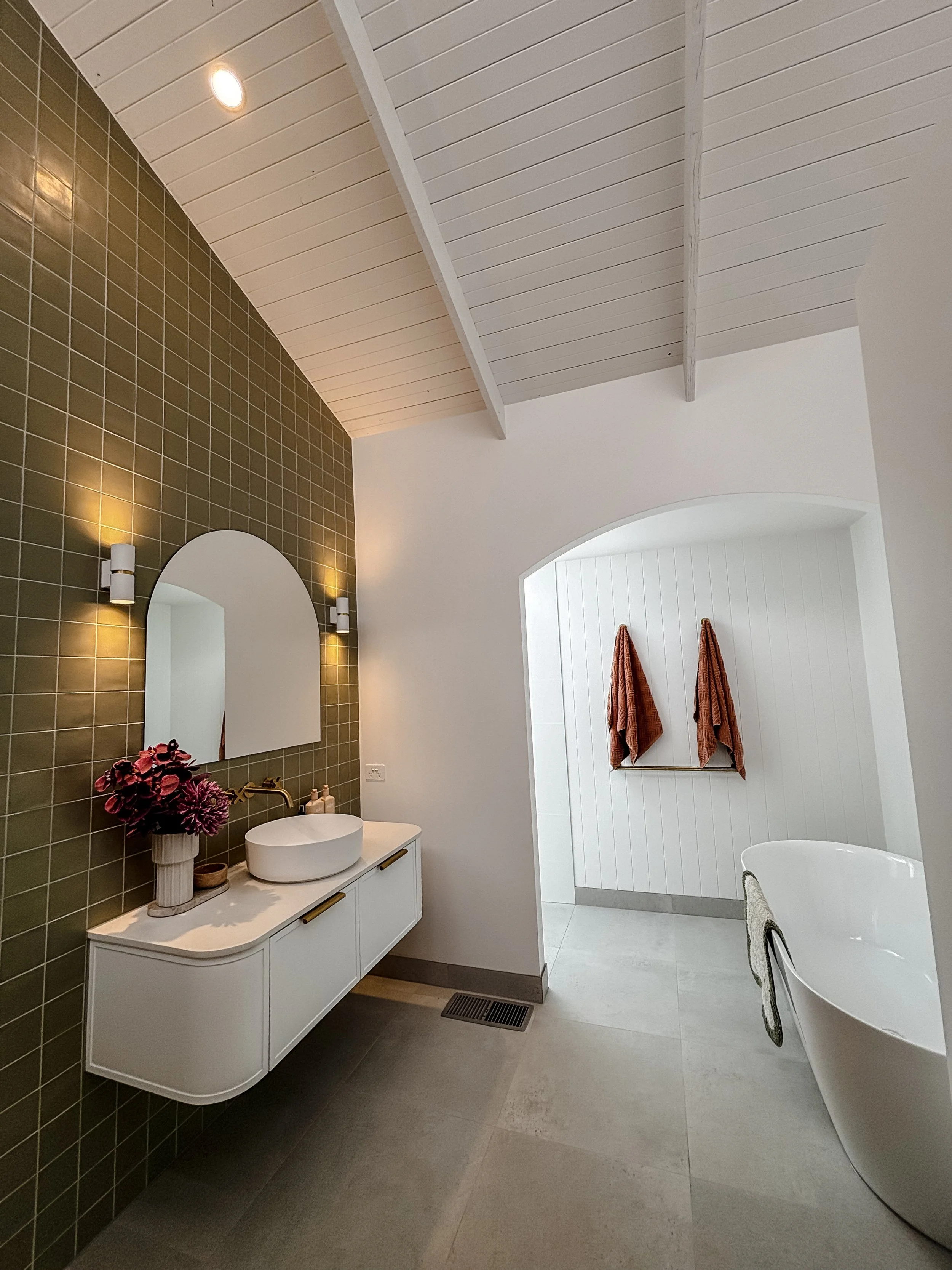 Modern bathroom with white vanity and round vessel sink, green tiled wall, arched mirror, a vase with purple flowers, two wall sconces, a towel rack with orange towels, a white bathtub, and a ceiling with white painted wooden beams.