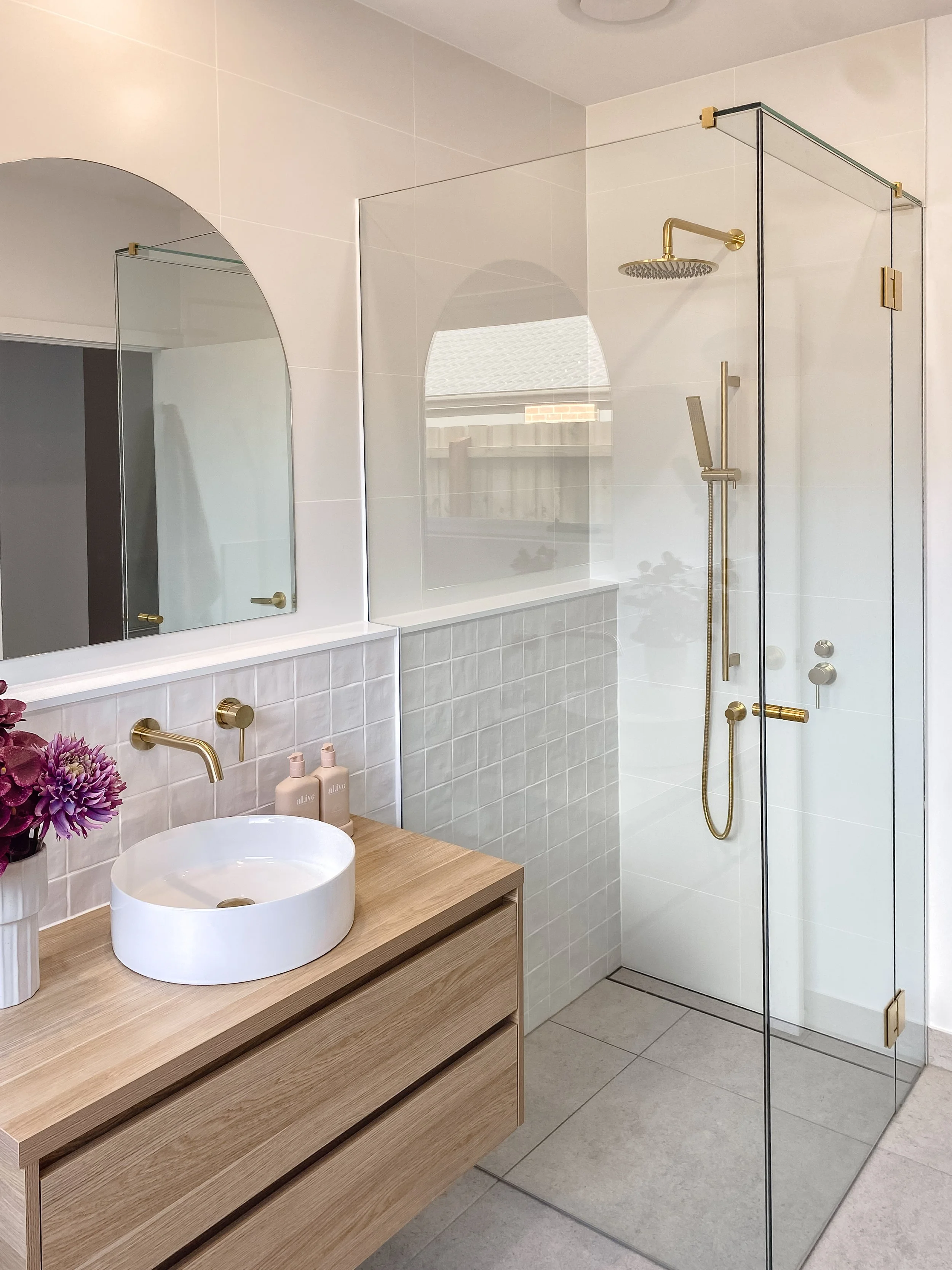 A modern bathroom featuring a wooden vanity with a round white vessel sink, a wall-mounted gold faucet, and a large mirror. There are bottles of lotion and a vase with purple flowers on the vanity. The room includes a tiled shower with a glass enclosure, a gold showerhead, and a handheld showerhead, with a floor drain visible.