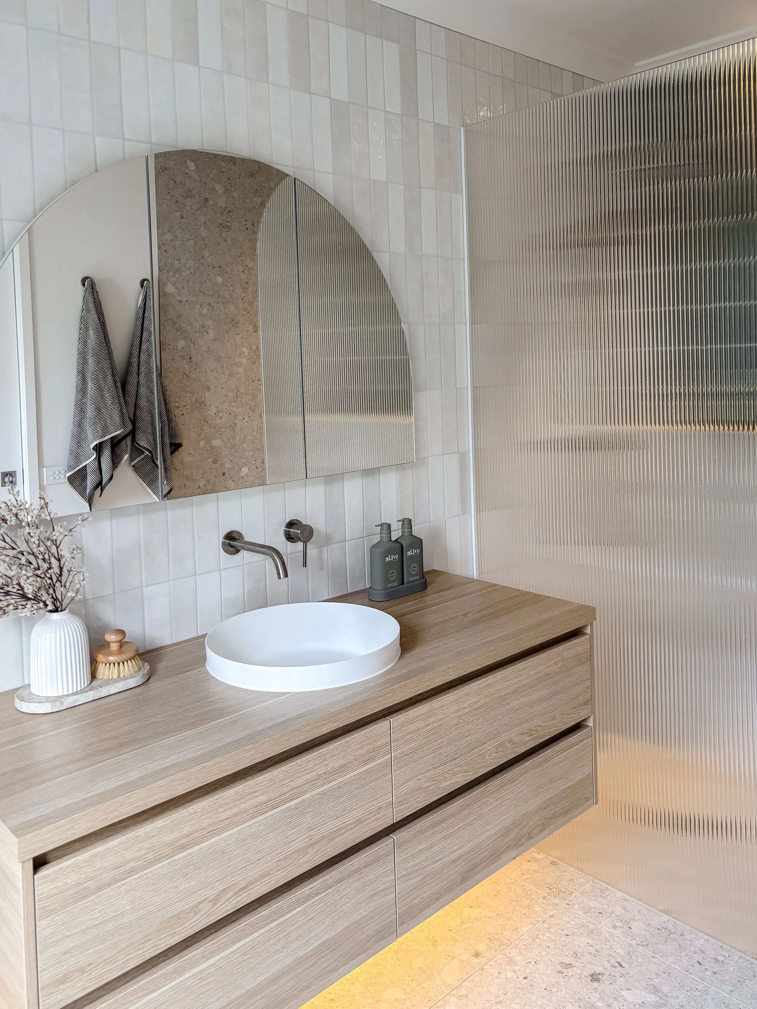Modern bathroom with a wooden vanity, a round white vessel sink, a wall mirror, two striped towels hanging, bottles of soap, a decorative vase, and a textured metallic sliding door. The walls are tiled with white tiles and the floor is beige.