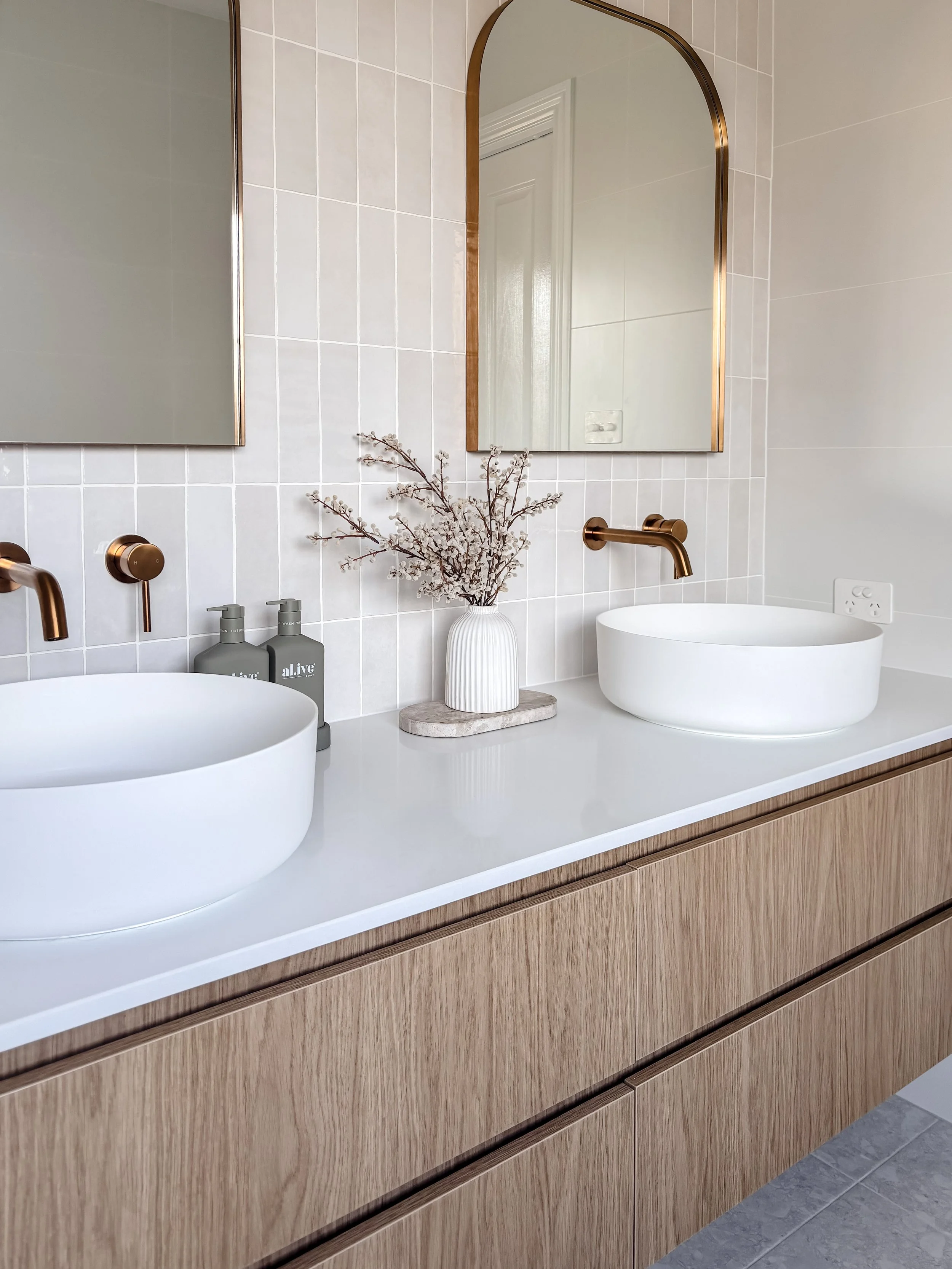 A modern bathroom vanity with two white vessel sinks, a vase with dried flowers, and two soap dispensers. The vanity has a light wood finish with sleek drawers, and the backsplash features white tiles. There are two mirrors with gold frames above each sink, and a single gold spout faucet for each basin. An electrical outlet is visible on the wall.