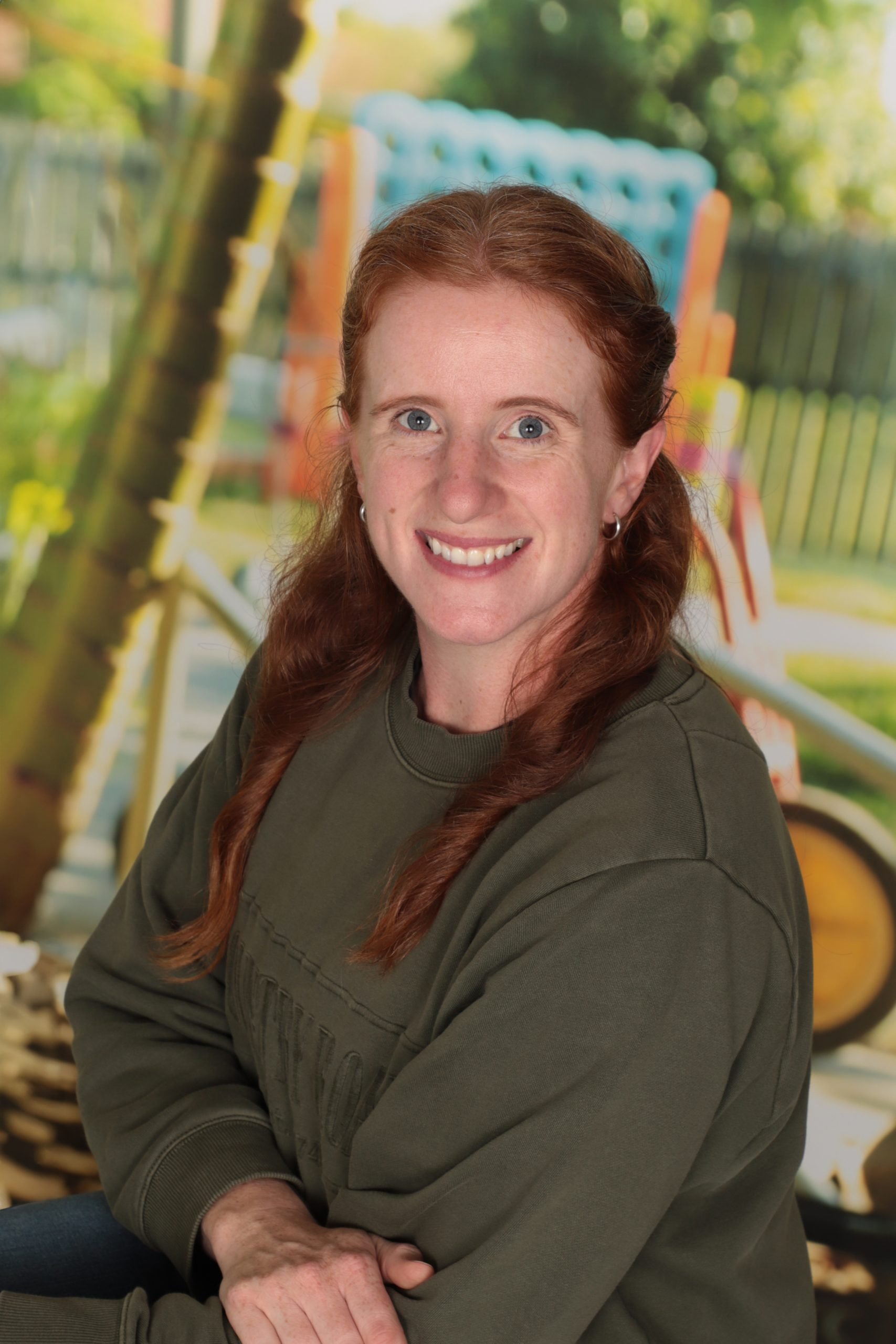 A woman with red hair and blue eyes wearing a dark green jacket, sitting outdoors with a colorful park or playground in the background.
