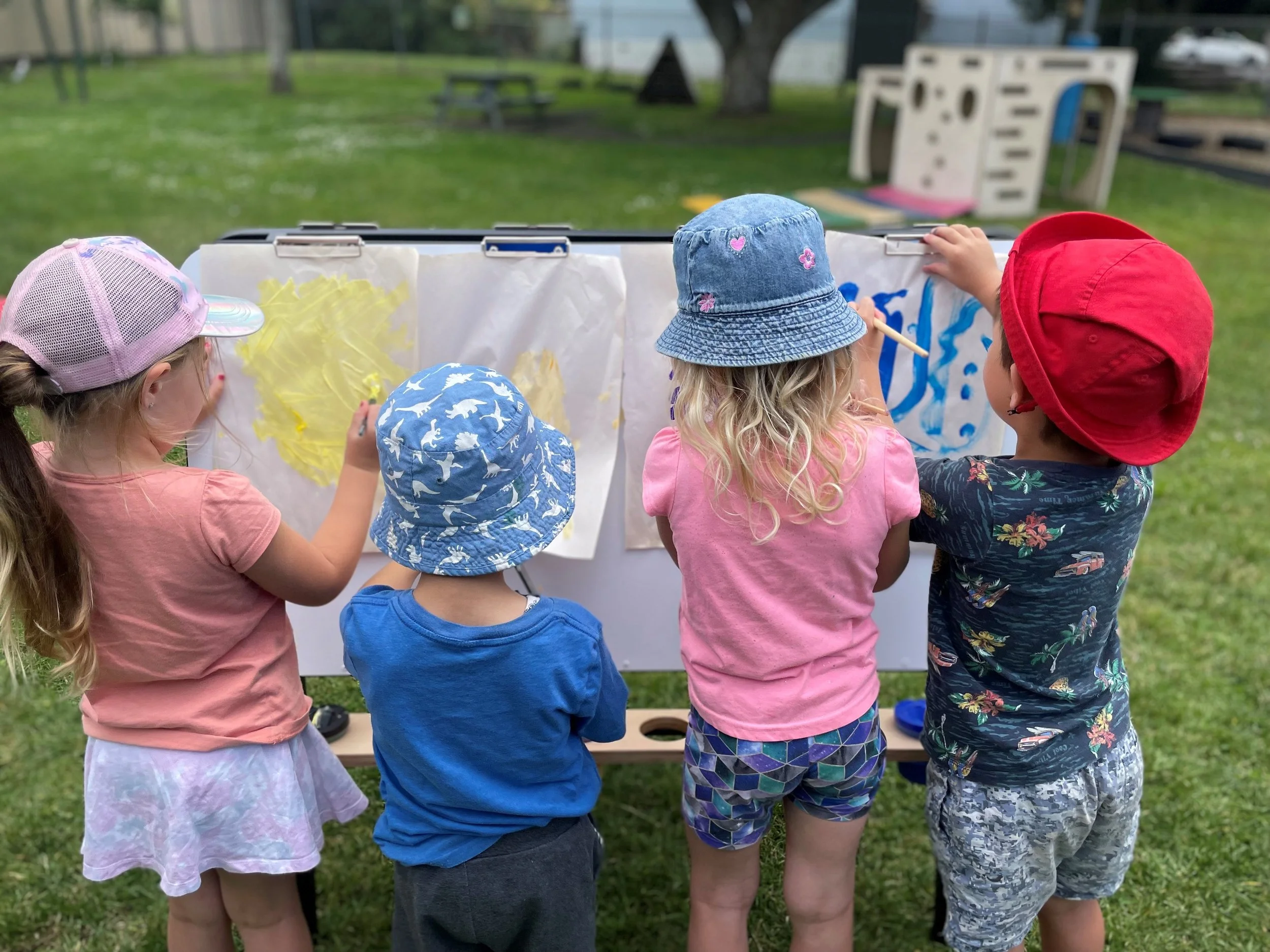 Children wearing colorful hats and shorts painting on large white paper outdoors, with a grassy area and playground equipment in the background.
