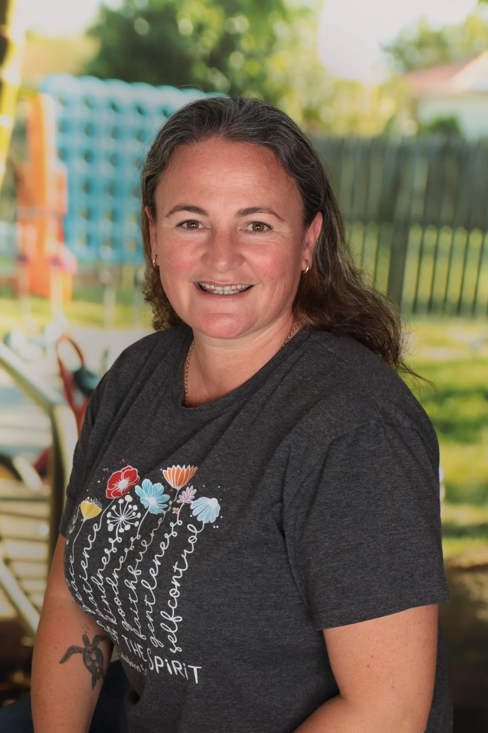 A smiling woman with long brown hair, wearing earrings and a dark gray t-shirt with colorful floral design and text, sitting outdoors with a blurred background of trees and a fence.