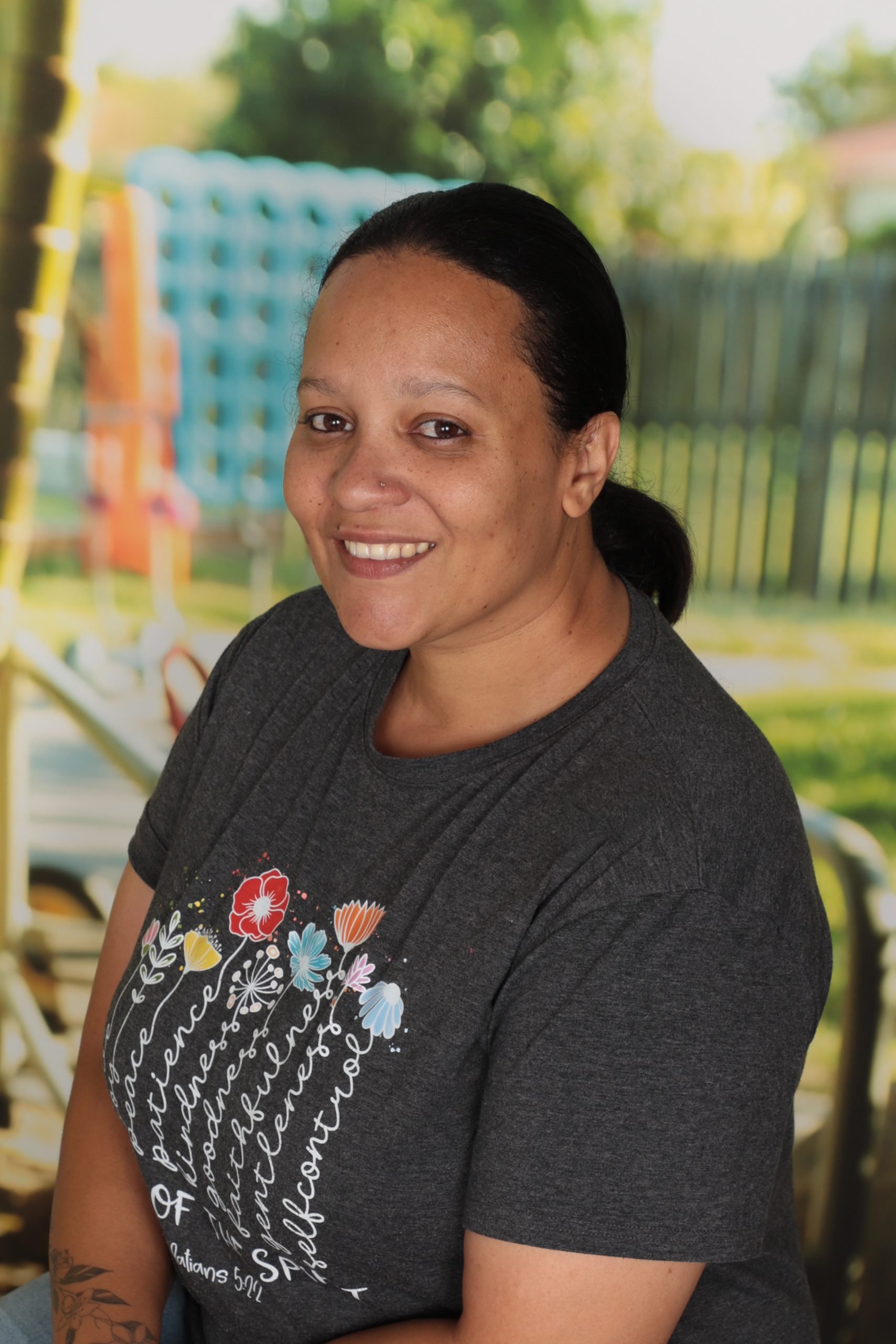 A smiling woman with dark hair pulled back, wearing a dark gray shirt with colorful floral and script design, in an outdoor setting with a blurred background of trees and a playground.
