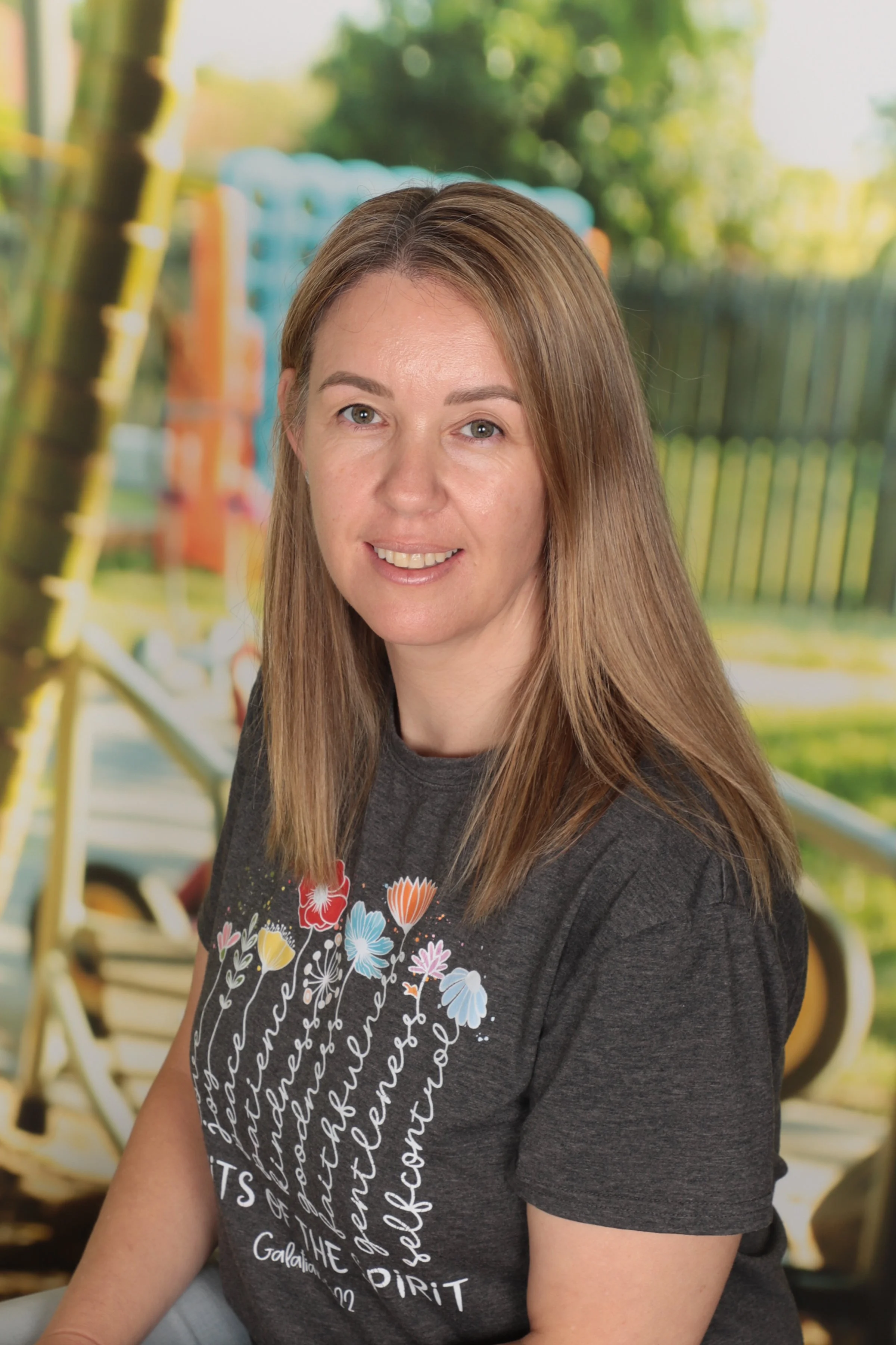 A woman with long, straight brown hair and light skin, smiling at the camera. She is wearing a dark gray t-shirt with colorful floral and script design. The background features a blurred outdoor scene with green trees and playground equipment.