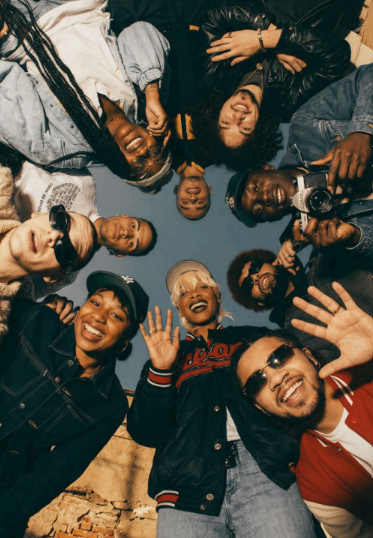 Group of diverse friends lying in a circle on the ground, smiling and waving at the camera indoors.