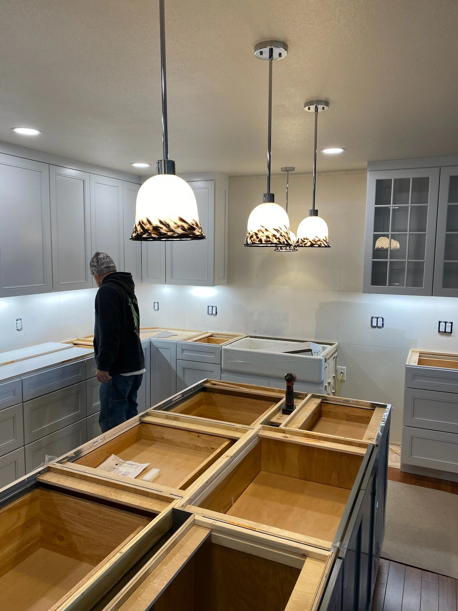 Kitchen cabinetry being installed, with a worker present, and pendant lights hanging from the ceiling.