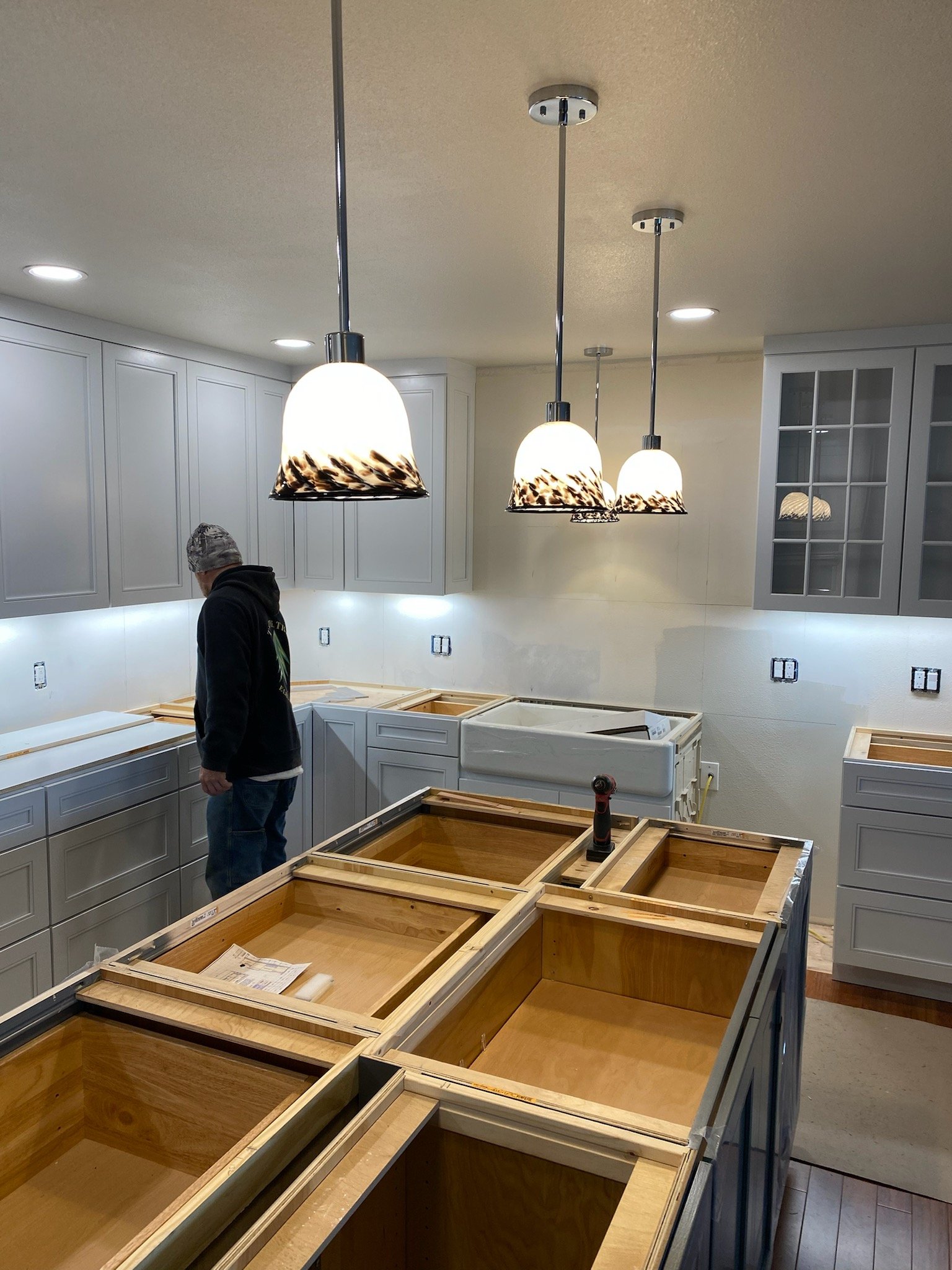Kitchen with white cabinets, three hanging pendant lights, and an ongoing cabinet installation with empty wooden frames on countertops and some tools. A person is working.