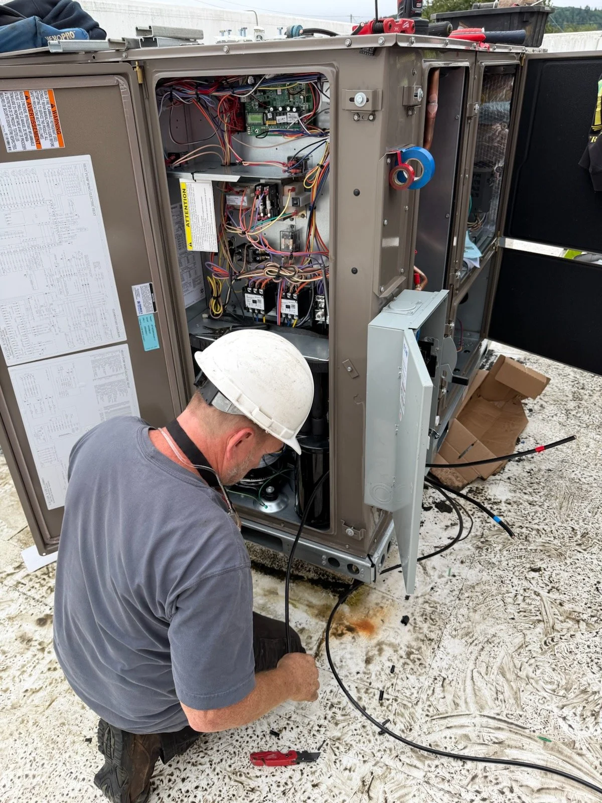A technician wearing a hard hat working on the electrical components of an outdoor HVAC or industrial equipment unit, with wiring and internal parts visible.