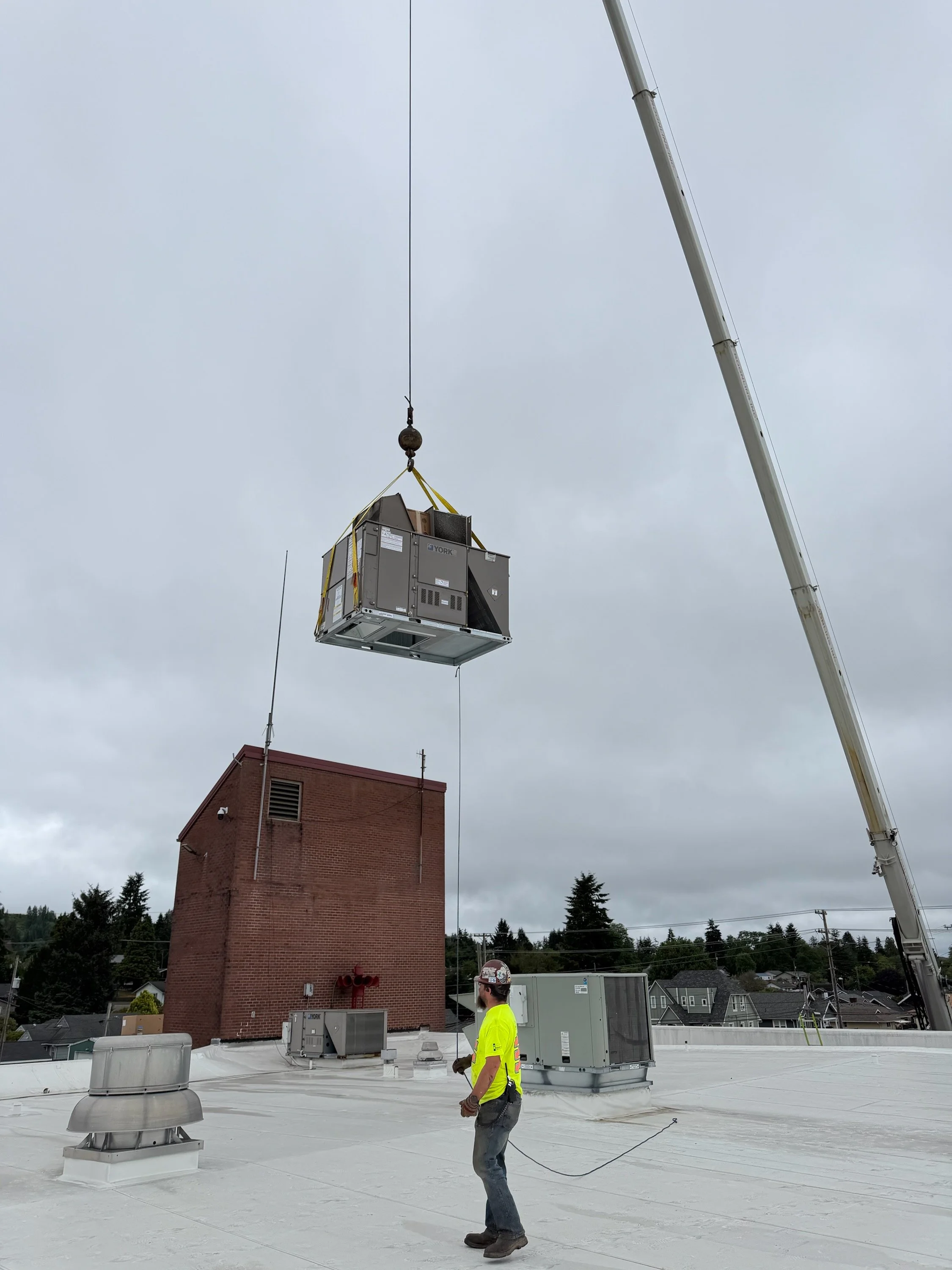 A construction worker wearing a yellow safety vest and a hard hat on a flat rooftop, overseeing the lifting of a large HVAC unit by a crane through a hanging cable, with an overcast sky in the background.