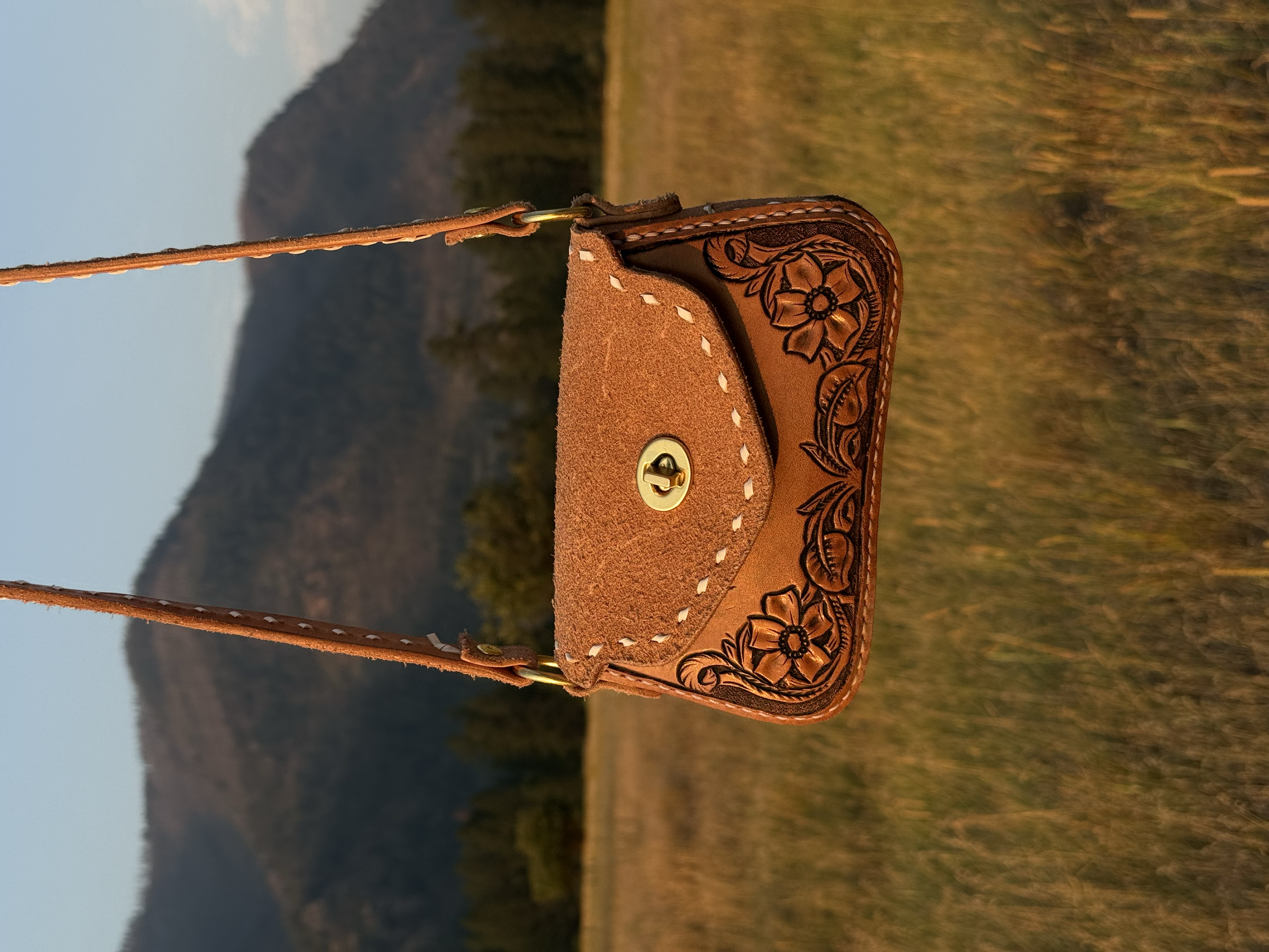 A close-up of a leather saddlebag with floral tooling and stitching, hanging on a strap with a metal clasp, set against a landscape with grassy fields and hills in the background during sunset.