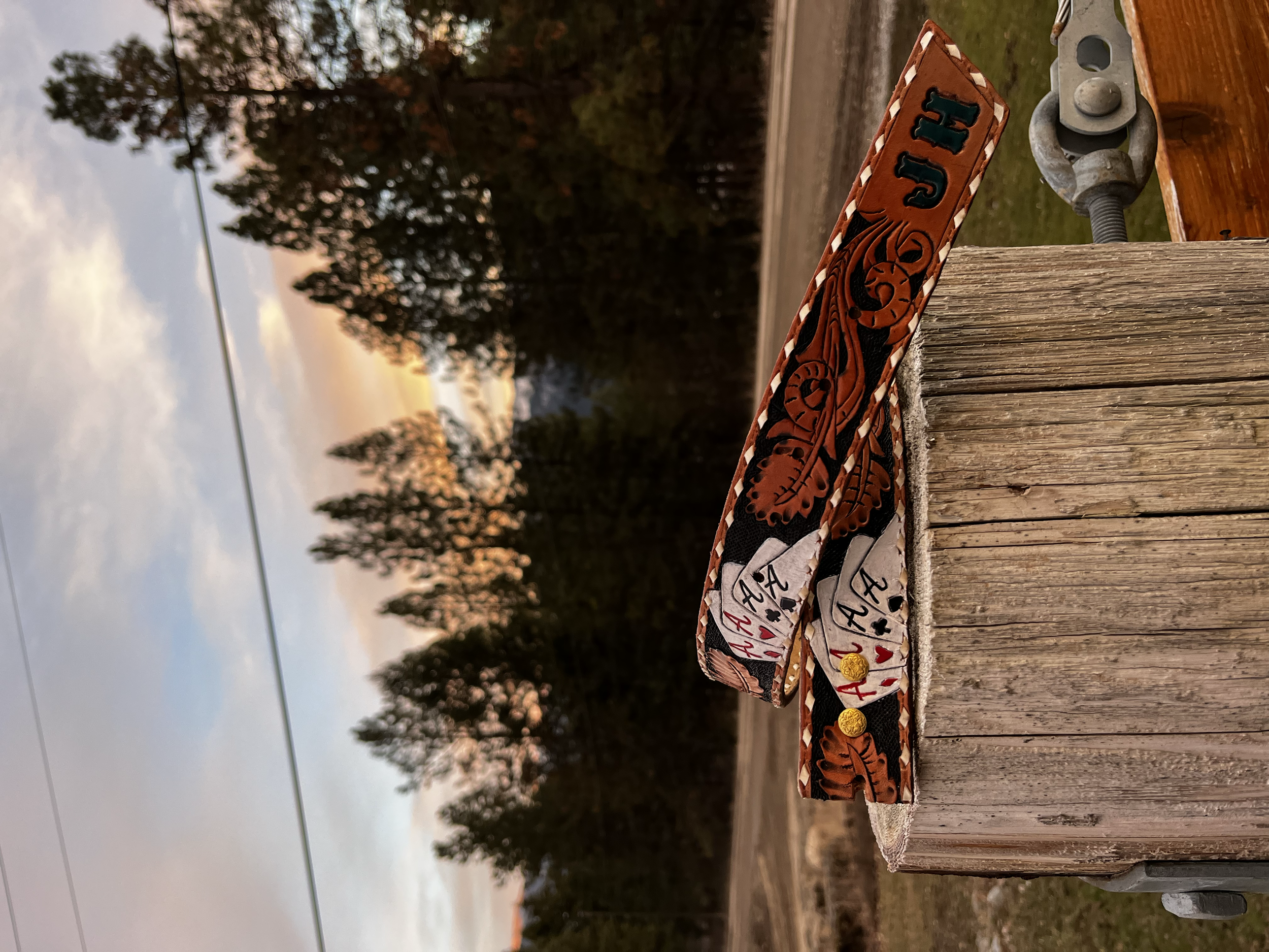 A leather belt with playing card suits and numbers, hanging off a wooden post outdoors during sunset.