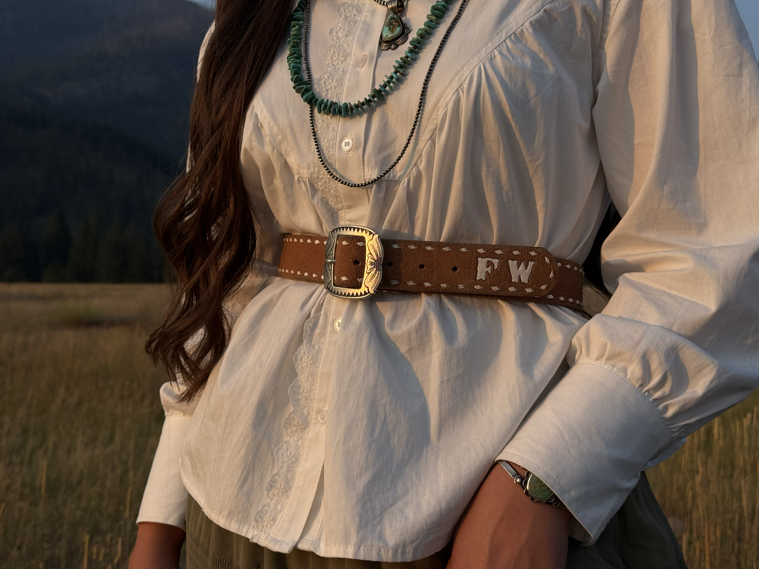 Close-up of a woman wearing a white embroidered blouse with layered necklaces, a wide brown leather belt with a detailed silver buckle, and a bracelet, standing outdoors at sunset or sunrise.