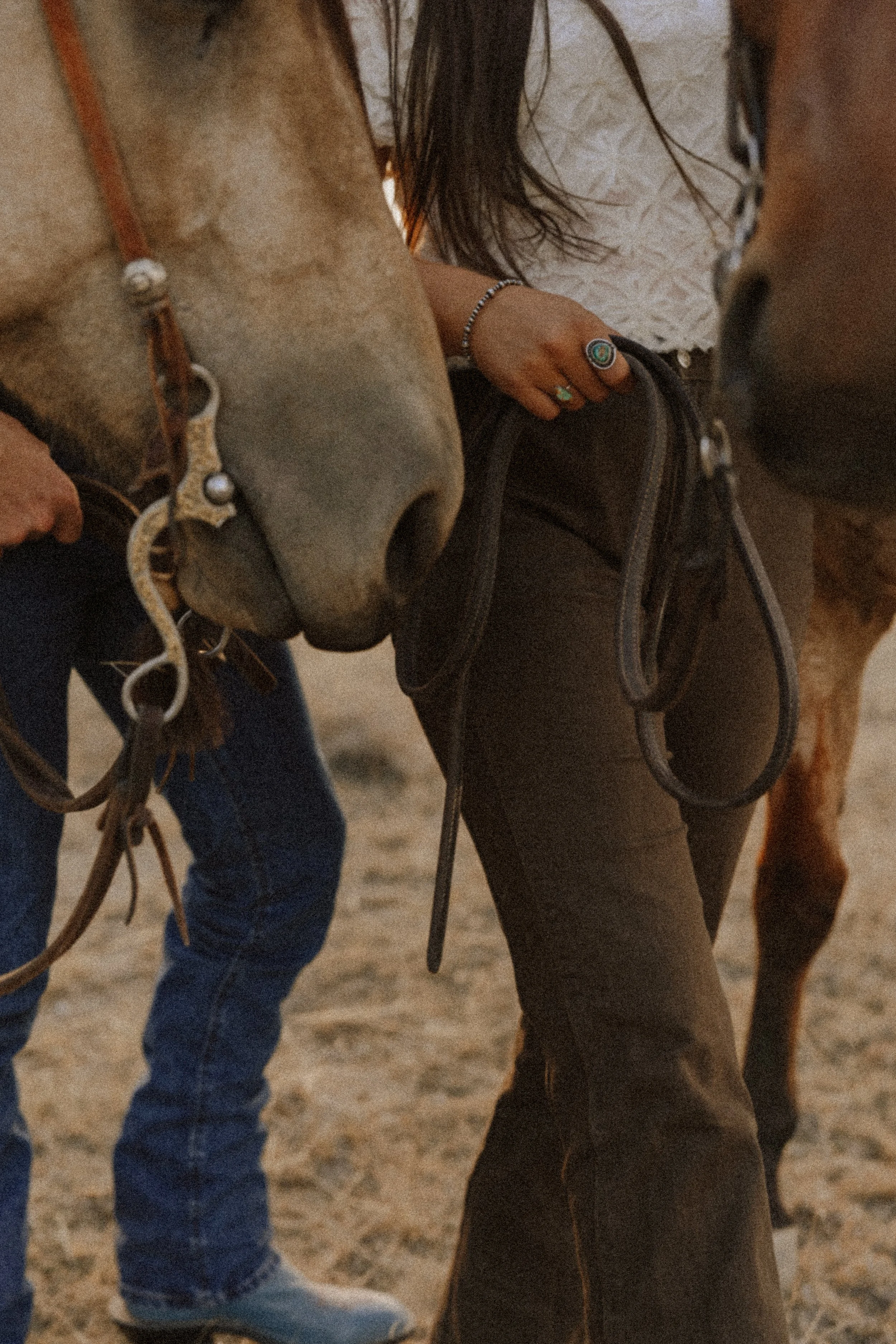 Close-up of two people preparing to ride horses, focusing on their hands and waist area, showing details of their clothing, accessories, and horse tack.