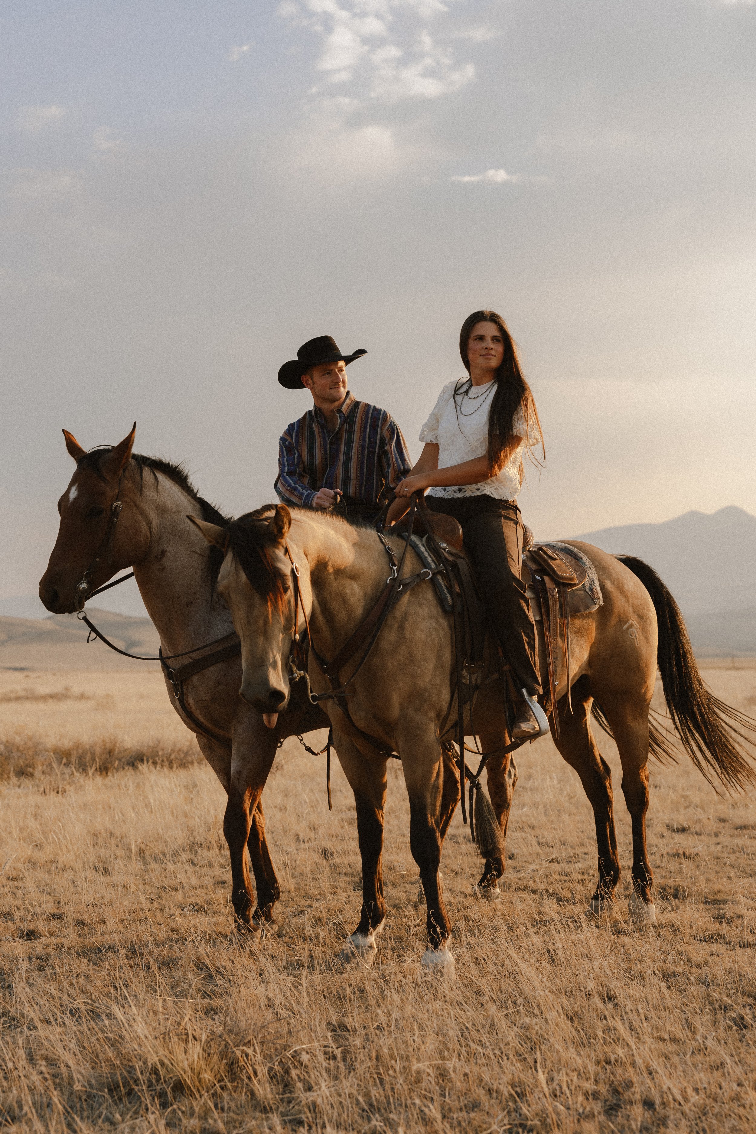 A man and a woman riding horses across a dry, grassy plain at sunset, with mountains in the background.