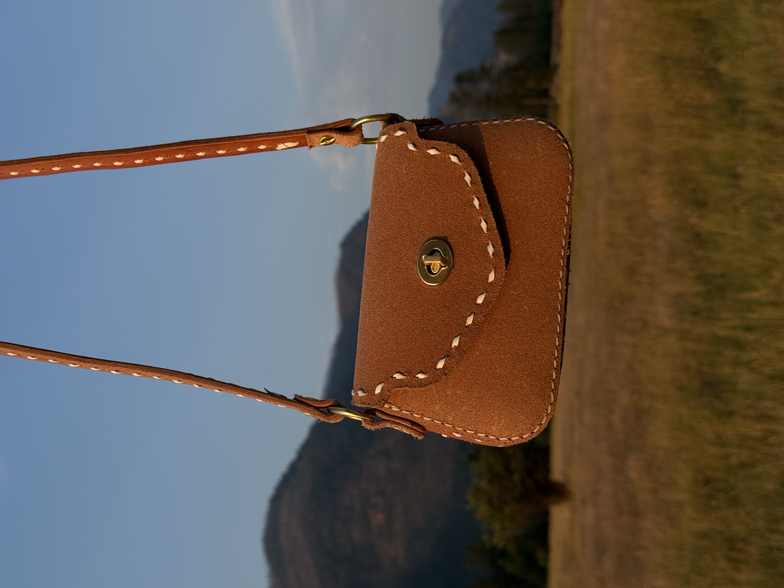 A brown leather handbag with white stitching and a gold clasp, hanging from a strap against a landscape background of mountains, fields, and a blue sky.