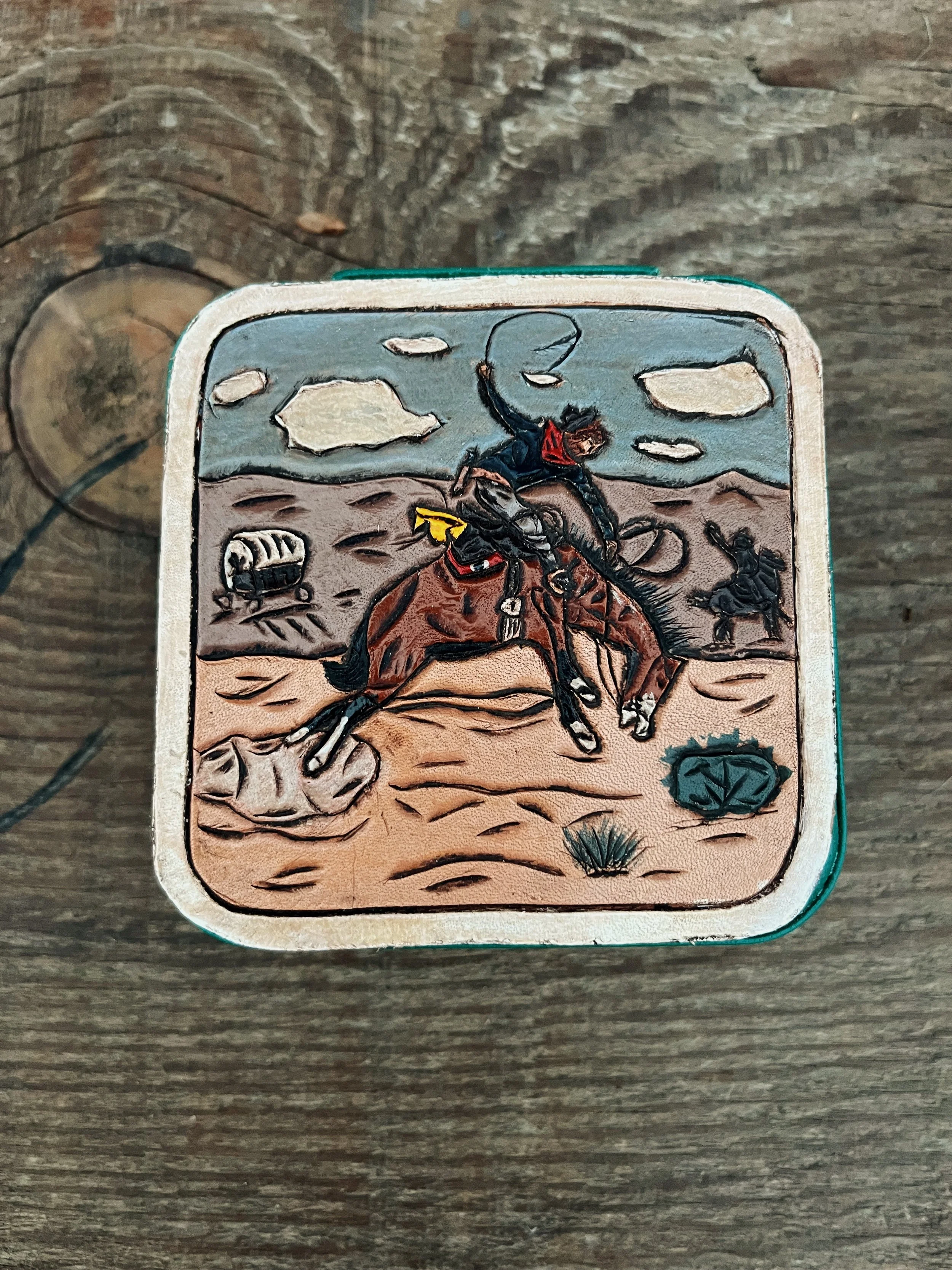 A painted illustration on a rectangular object depicts a cowboy riding a horse in a desert, with mountains and clouds in the background, a cactus, a tumbleweed, and a small shield symbol on the bottom right corner.