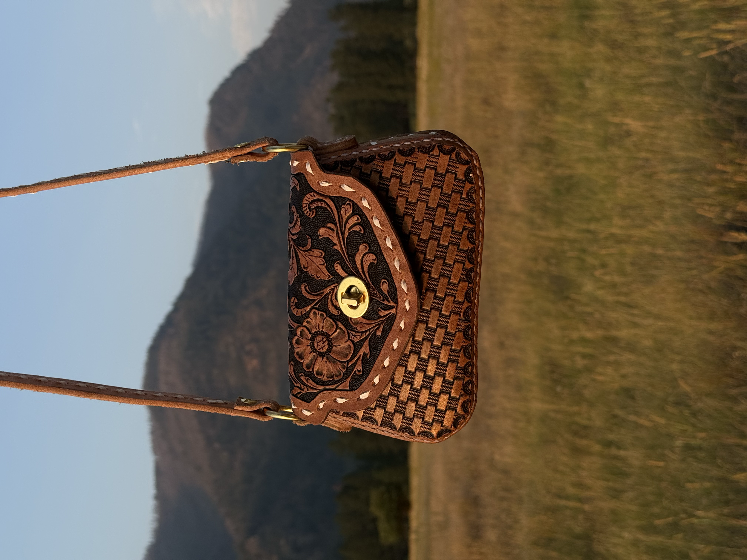 Close-up of a woven leather handbag with floral embroidery, gold clasp, and brown leather strap, hanging outdoors during sunset with mountains in the background.