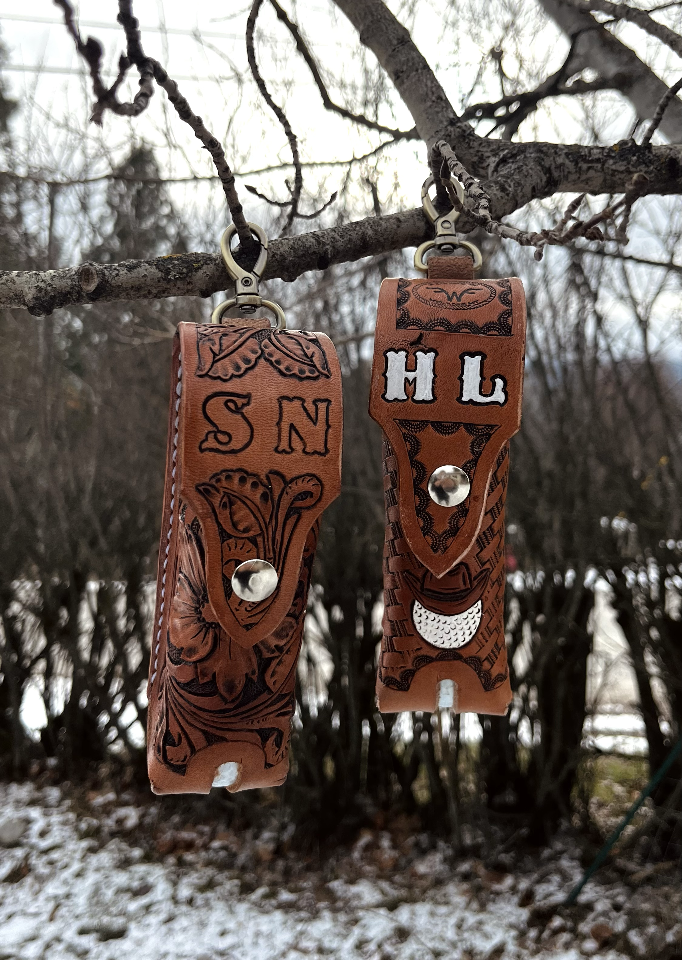 Close-up of two leather straps with engraved designs hanging from a tree branch, with a blurred outdoor background of leafless trees and snow-covered ground.