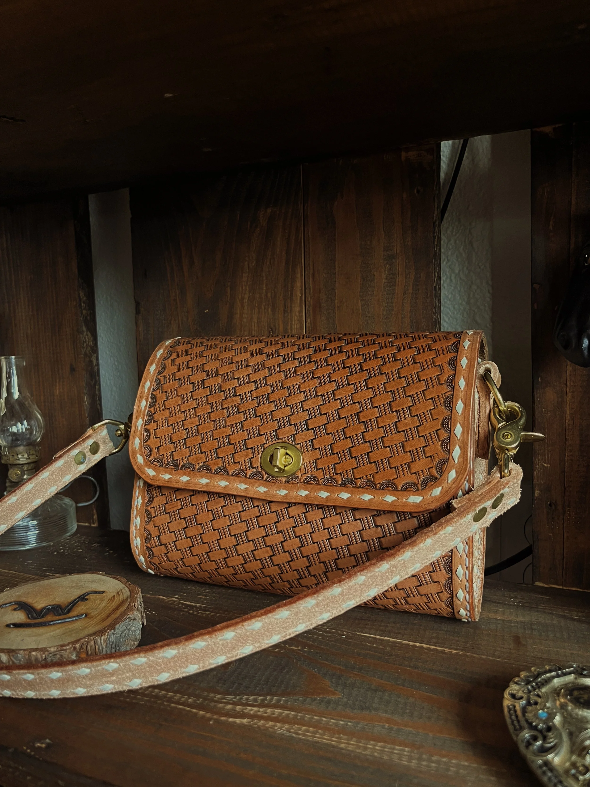 A woven leather purse with a gold clasp resting on a wooden surface, with a lamp and a wooden background partially visible.
