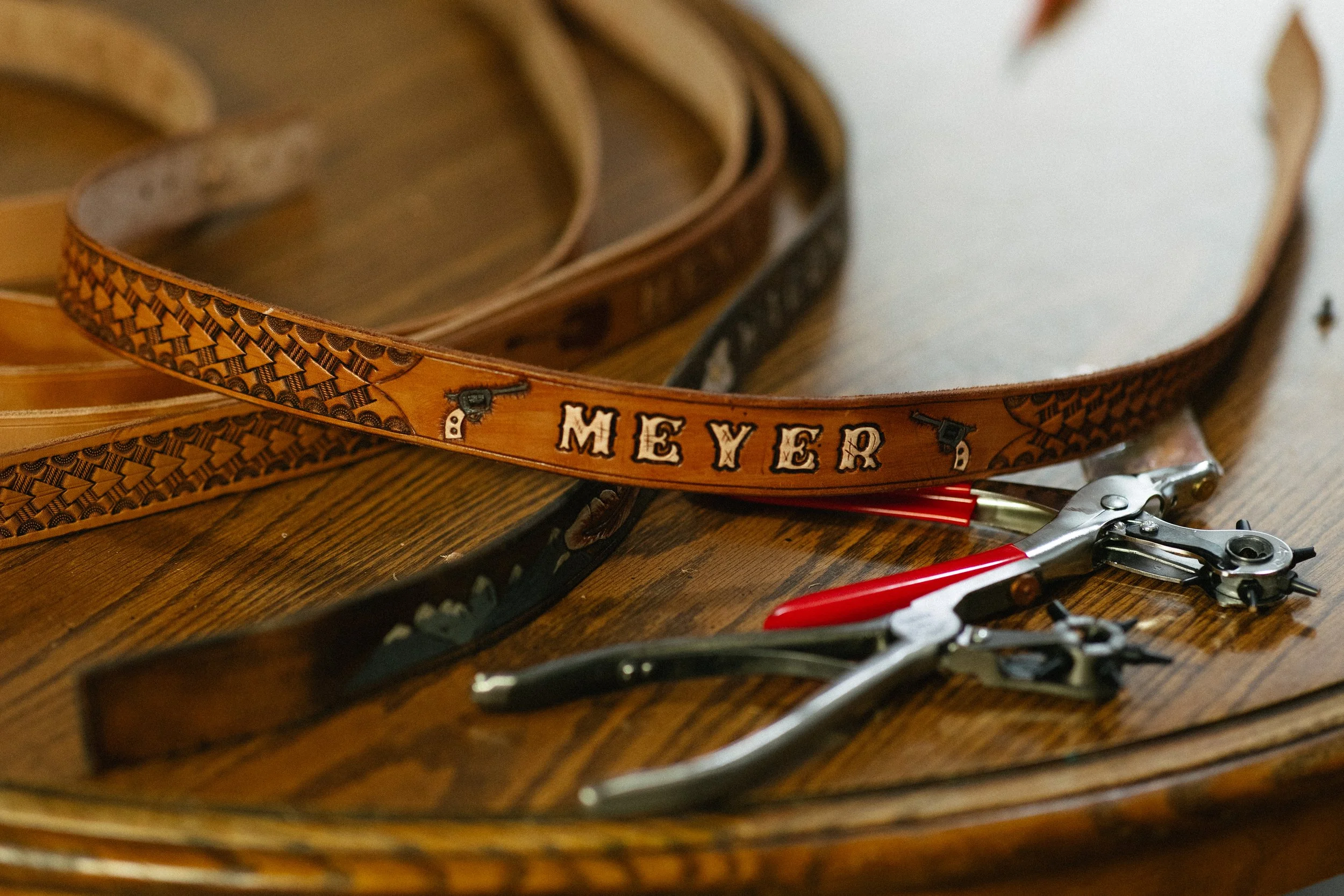 Close-up of a leather belt with orange and black patterns and the word 'MEXER' on it, along with other belts and tools on a wooden surface.