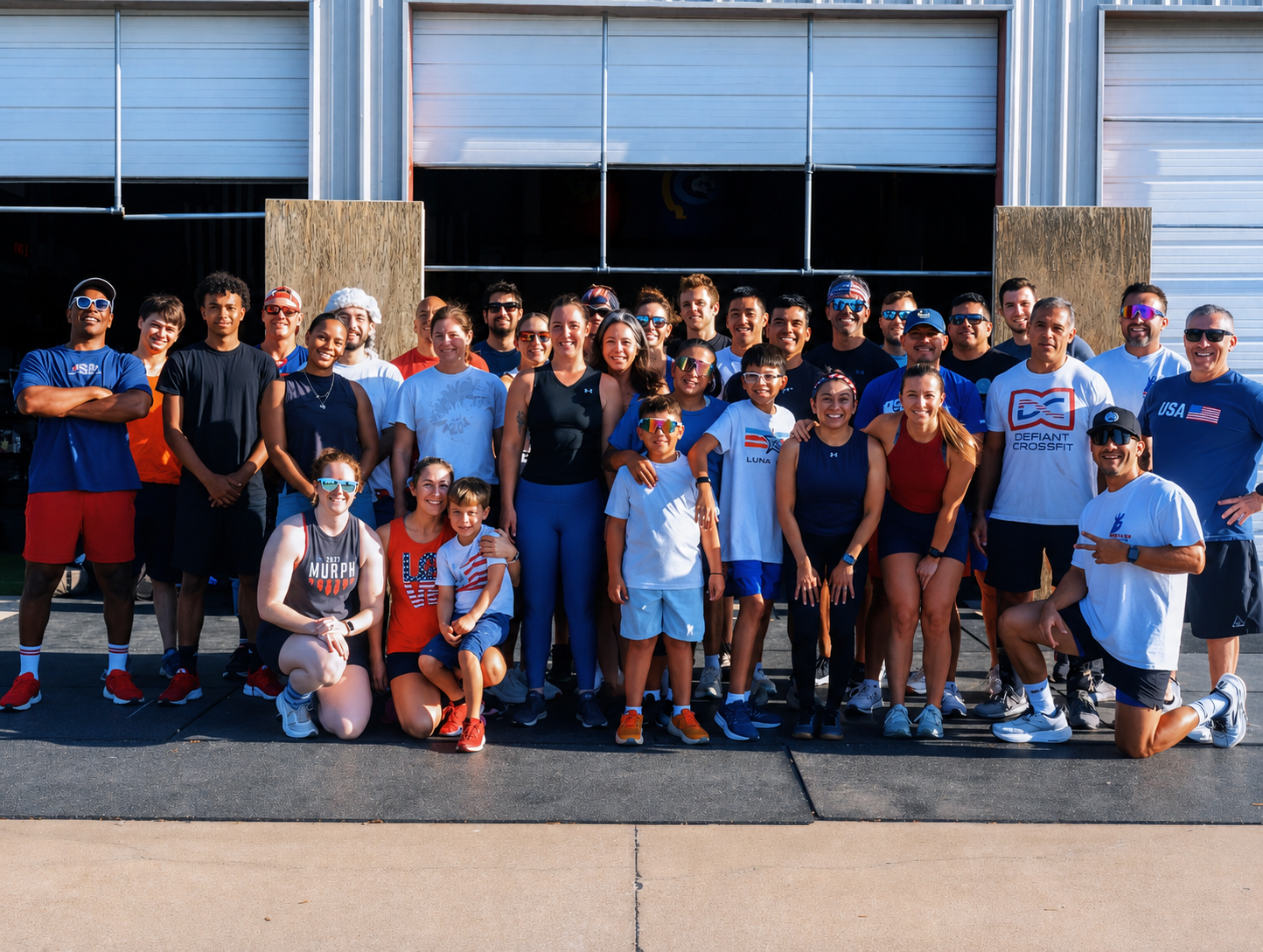 Group of diverse people, including children and adults, standing and kneeling in front of an industrial building, dressed in athletic clothing, sunglasses, and sporting CrossFit and USA-themed apparel, smiling at the camera during daytime.