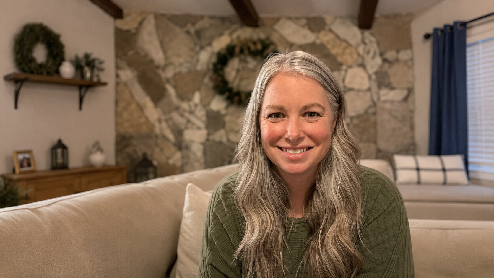 A smiling woman with long gray hair sitting on a beige couch in a cozy living room with a stone accent wall and blue curtains.