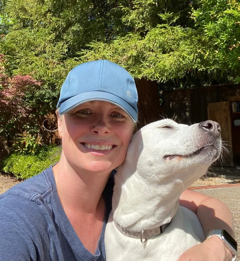 A smiling woman wearing a blue cap and a blue shirt, embracing a white dog with closed eyes and a content expression, outdoors in a sunny garden with green trees and bushes.