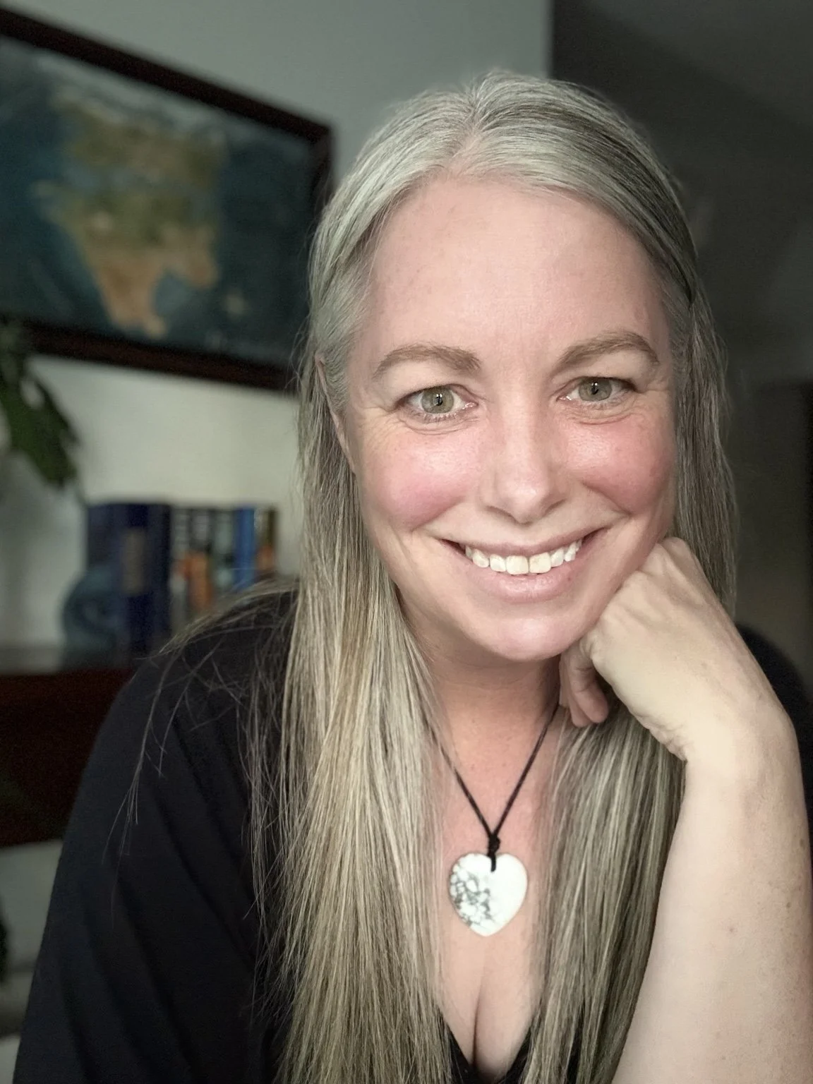 A woman with long gray hair smiling and resting her chin on her hand, wearing a black top and a heart-shaped pendant necklace. In the background, there is a blurred map of the United States on the wall and some books on a bookshelf.