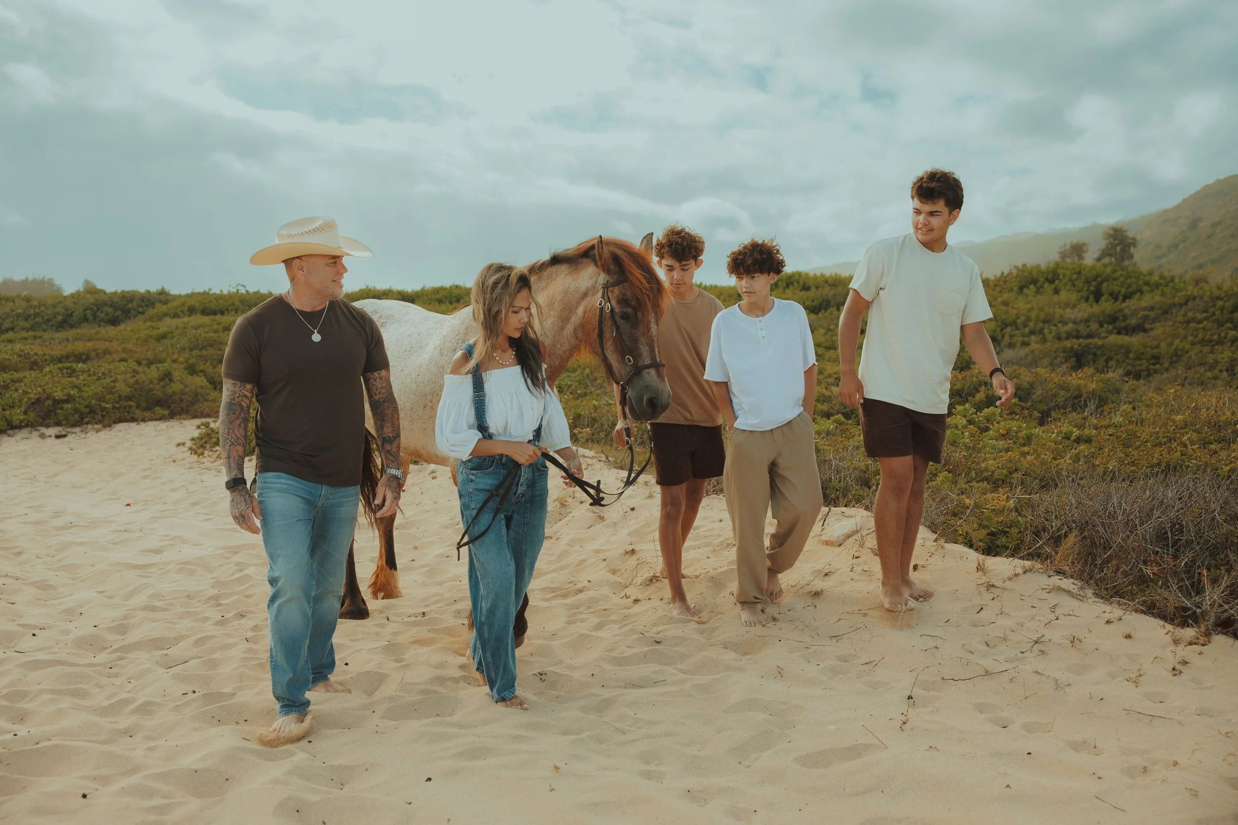 Group of five people walking with a horse on sandy terrain under a cloudy sky, with green bushes and hills in the background.