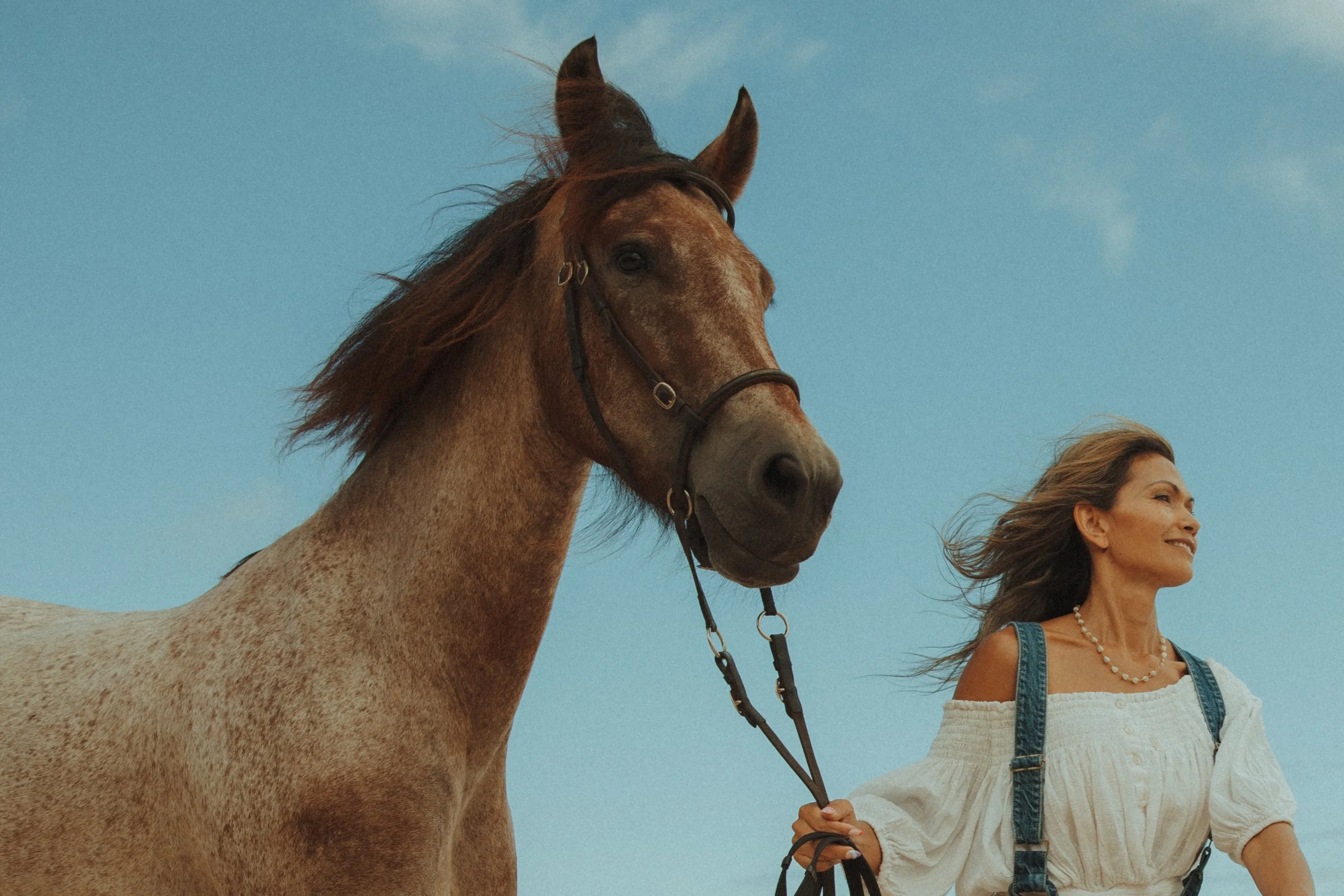 A woman with brown hair and a white off-shoulder top with denim straps walking a light brown horse on a lead against a blue sky.