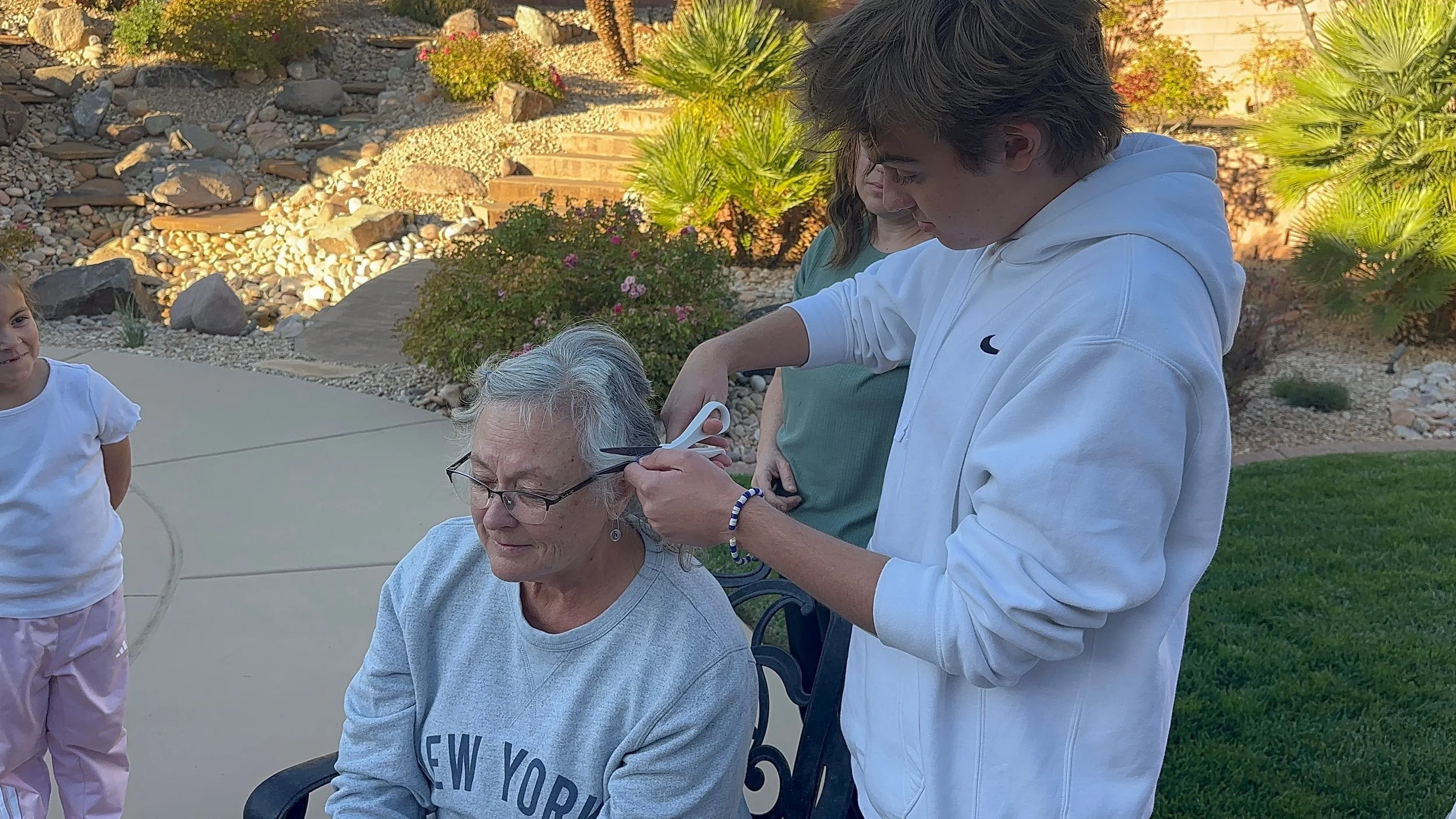 A young man is trimming the hair of an elderly woman seated outdoors, with two women and a girl nearby, in a landscaped backyard with rocks, plants, and flowers.