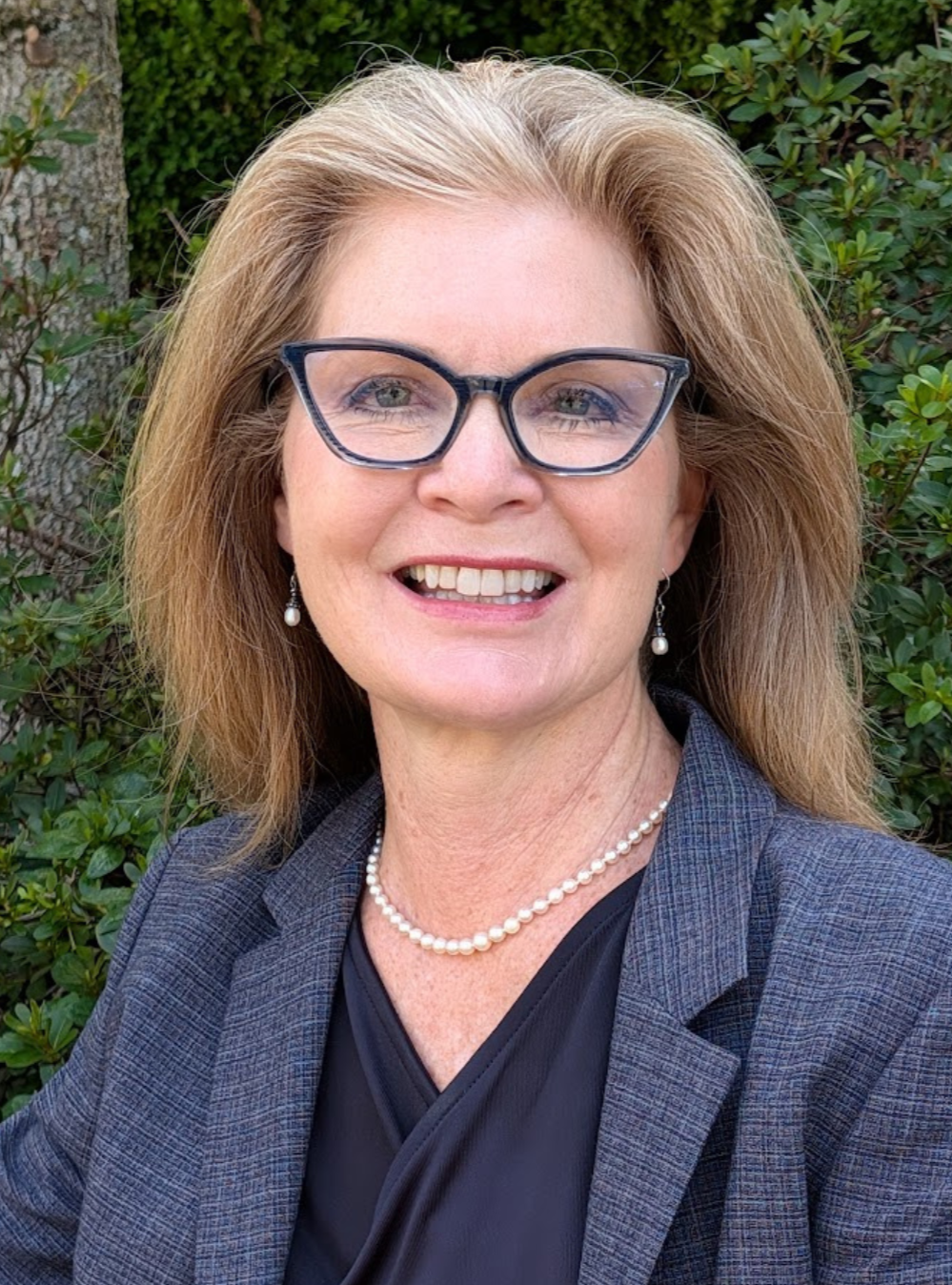 A smiling woman with blonde hair, wearing glasses, pearl earrings, a pearl necklace, a dark blouse, and a gray blazer, outdoors with green foliage in the background.
