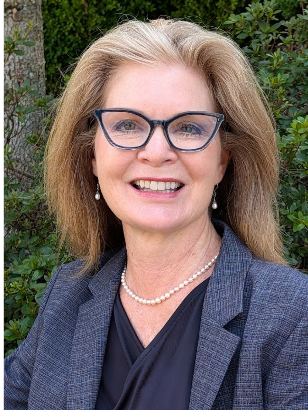 A smiling woman with light brown hair, wearing glasses, pearl earrings, a pearl necklace, a gray blazer, and a black top, standing outdoors with green bushes in the background.