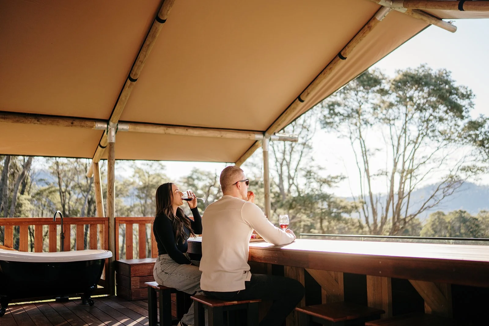 A man and a woman sitting at an outdoor bar drinking wine, with a mountain view and trees in the background under a shaded canopy.