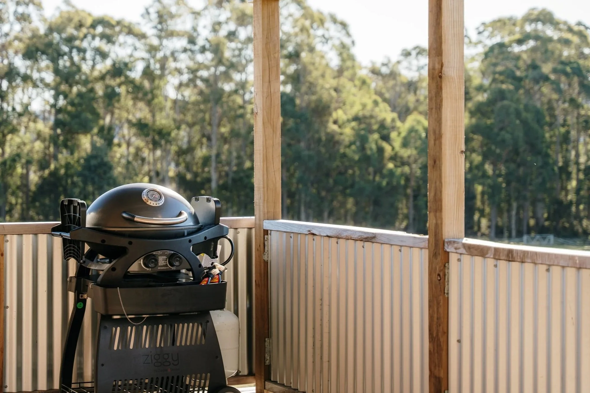 A black gas barbecue grill on a balcony with wooden posts and a metal railing, overlooking a wooded area in the background on a sunny day.