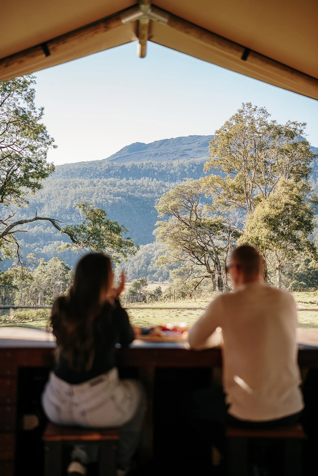 Two people sitting at a bar or counter looking out a large open window at a mountainous landscape with trees and a clear sky.