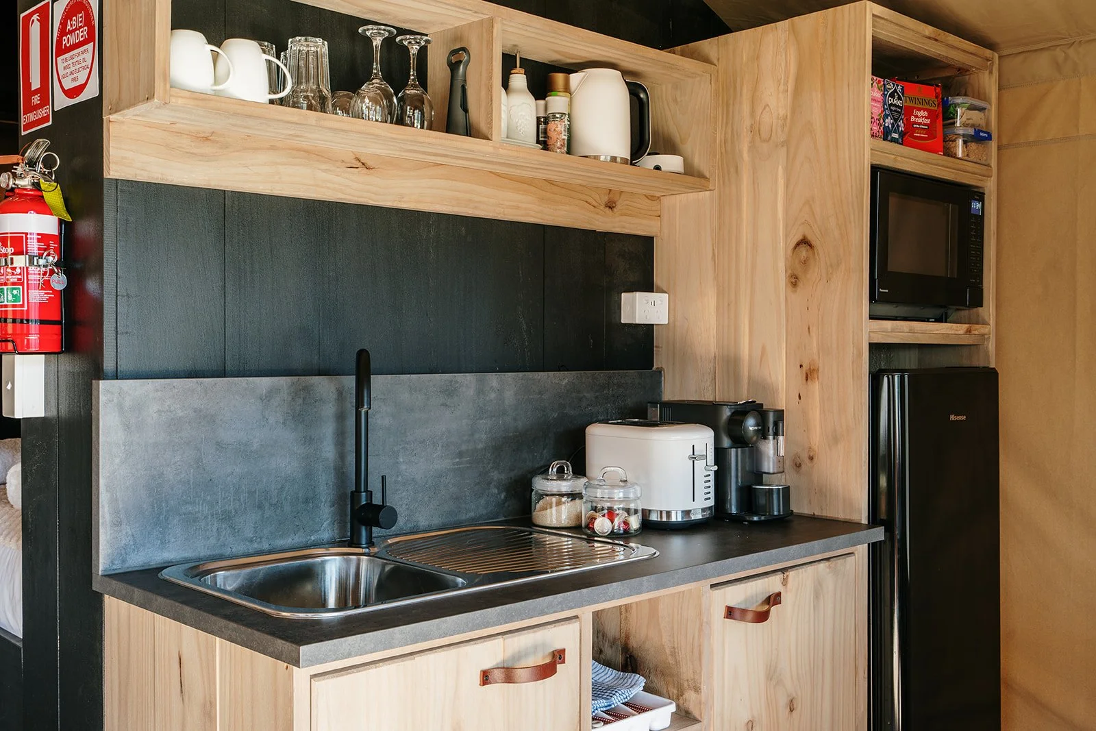 Kitchen with wood cabinetry, black countertop, gray backsplash, sink, coffee maker, rice cooker, microwave, mini refrigerator, and open shelves with glasses, cups, spices, and cereal boxes.