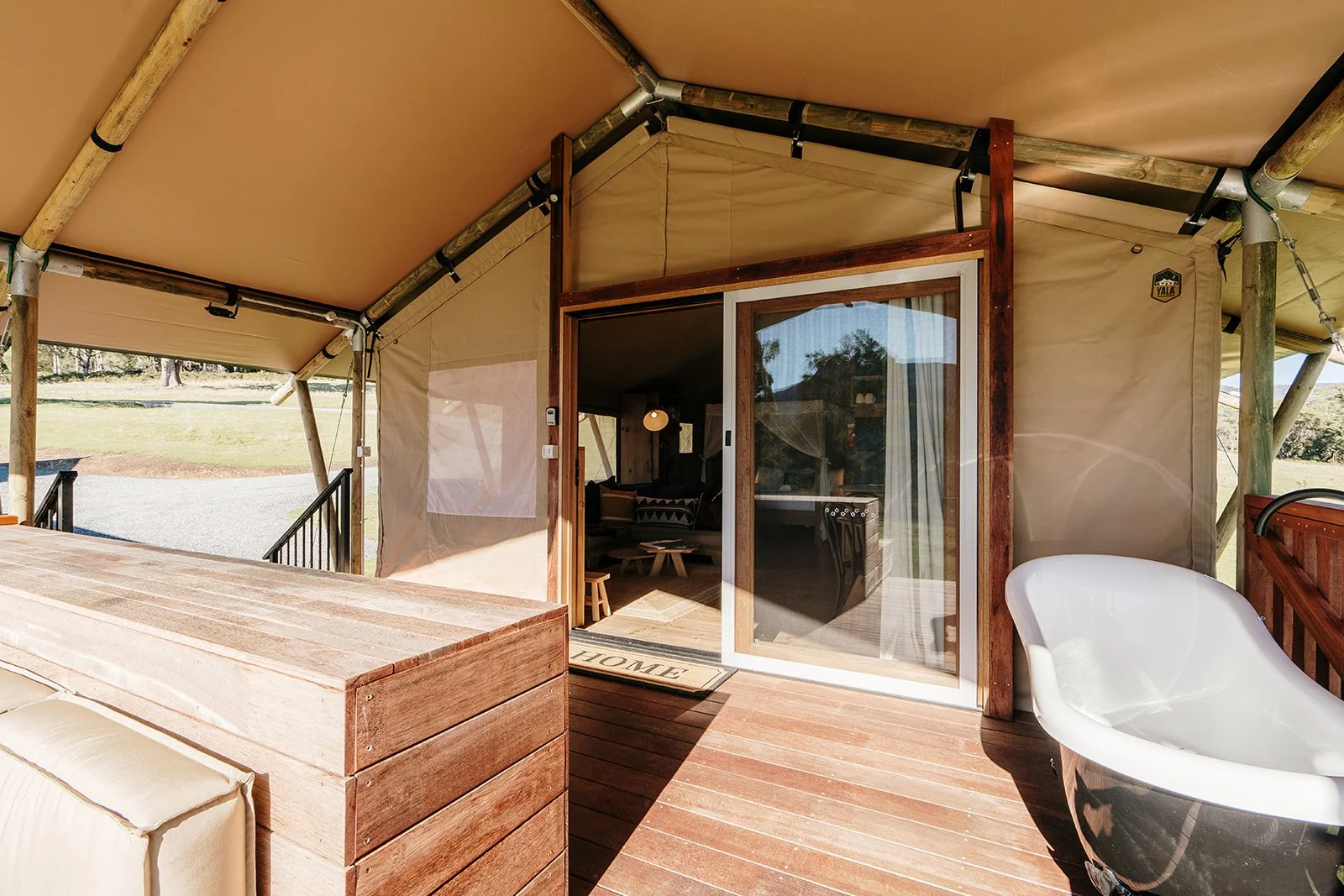 Outdoor patio area of a rustic tent cabin with wooden deck, sliding glass door, white bathtub, and beige canopy roof, surrounded by grassy landscape.
