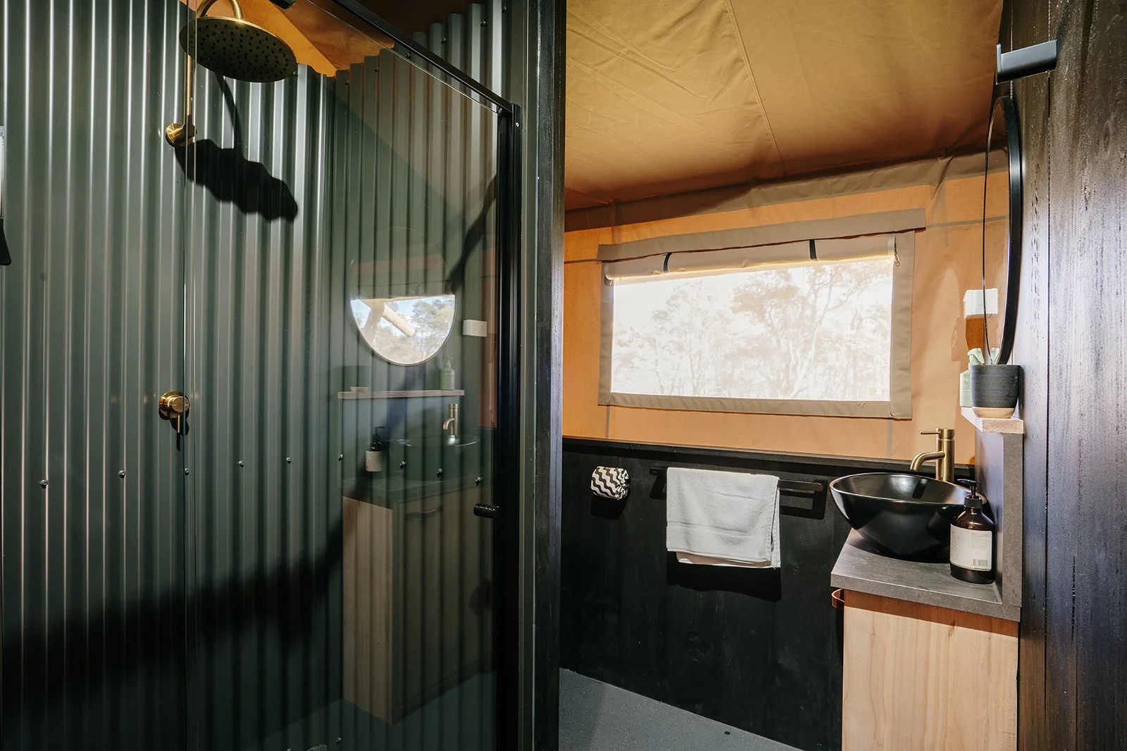 Modern bathroom with a glass shower, black vessel sink, brass fixtures, white towel, and natural light from a large window.
