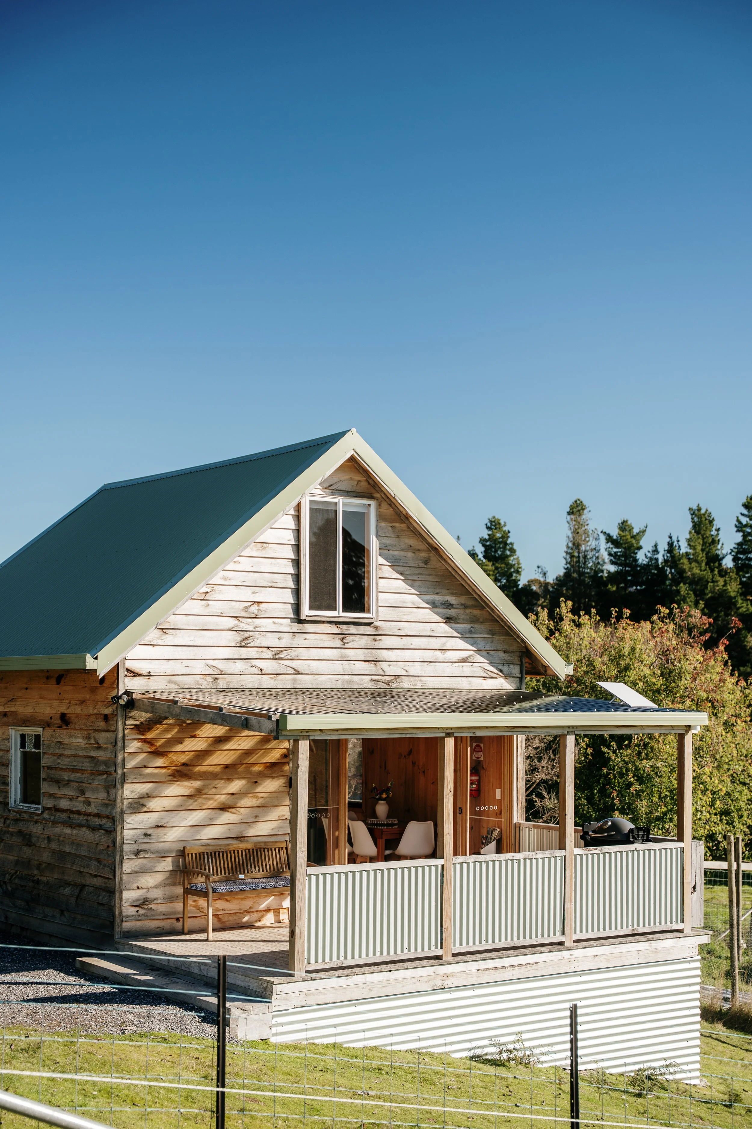 A wooden house with a gabled roof and a screened porch area, situated outdoors with trees in the background and a clear blue sky.