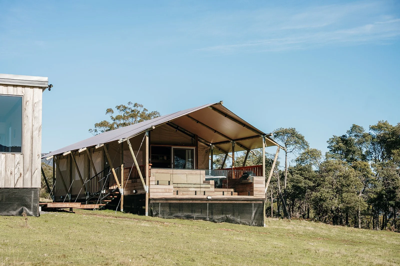 A house with a sloped roof and large deck, elevated on supports, surrounded by green grass and trees, under a clear blue sky.