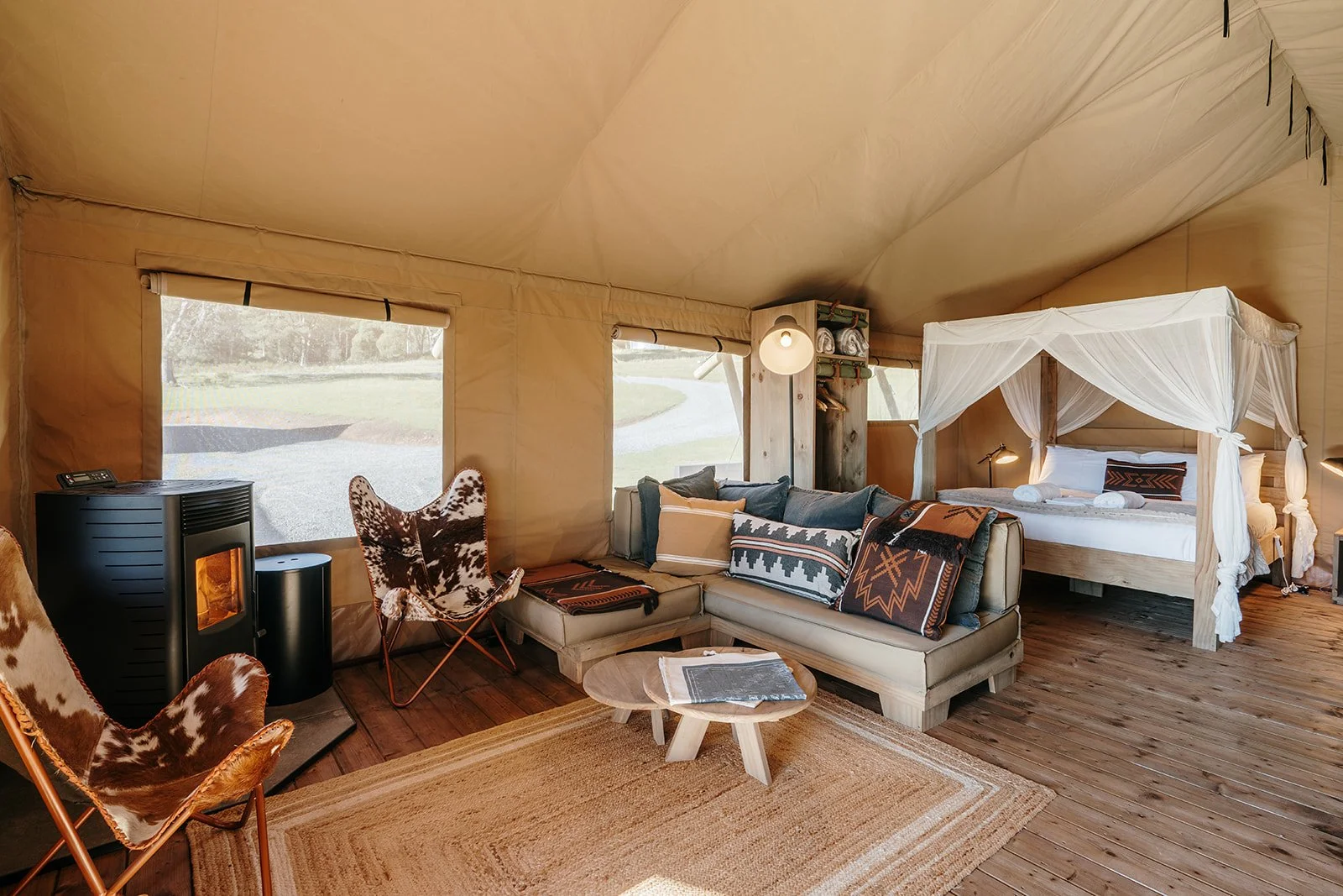 Interior of a luxury glamping tent with wooden floors, a four-poster bed with white canopy, a beige sofa with patterned pillows, cowboy boots-patterned armchair, small round wooden coffee table with magazines, a wood-burning stove, and large windows overlooking a grassy outdoor area.